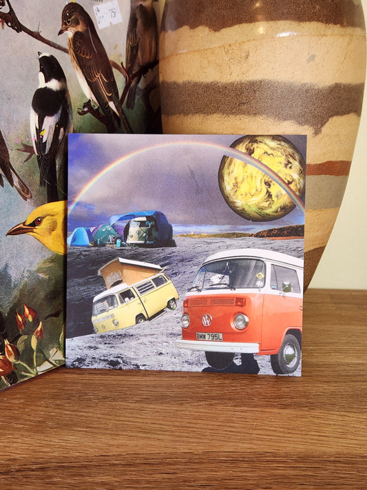 A square postcard featuring a surreal collage of vintage VW camper vans, a rainbow, and a planet, displayed on a wooden surface next to a large ceramic vase.