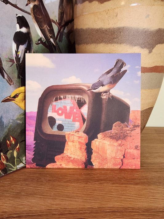A square postcard featuring a surreal collage of a television set with a bird perched on it and orange rock formations, displayed on a wooden surface next to a striped vase.