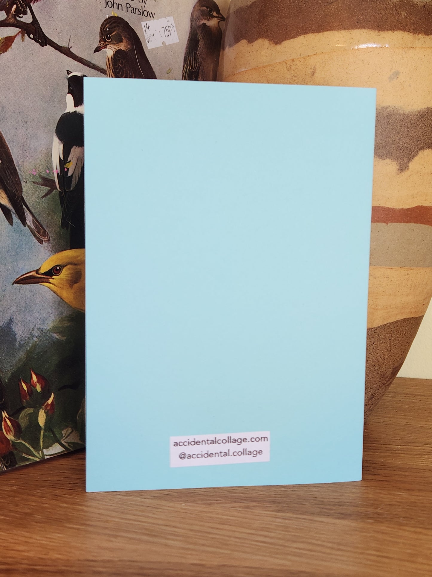 The back of a light blue greeting card on top of a wooden surface, with a small white label on the bottom reading 'accidentalcollage.com' and '@accidental.collage'.