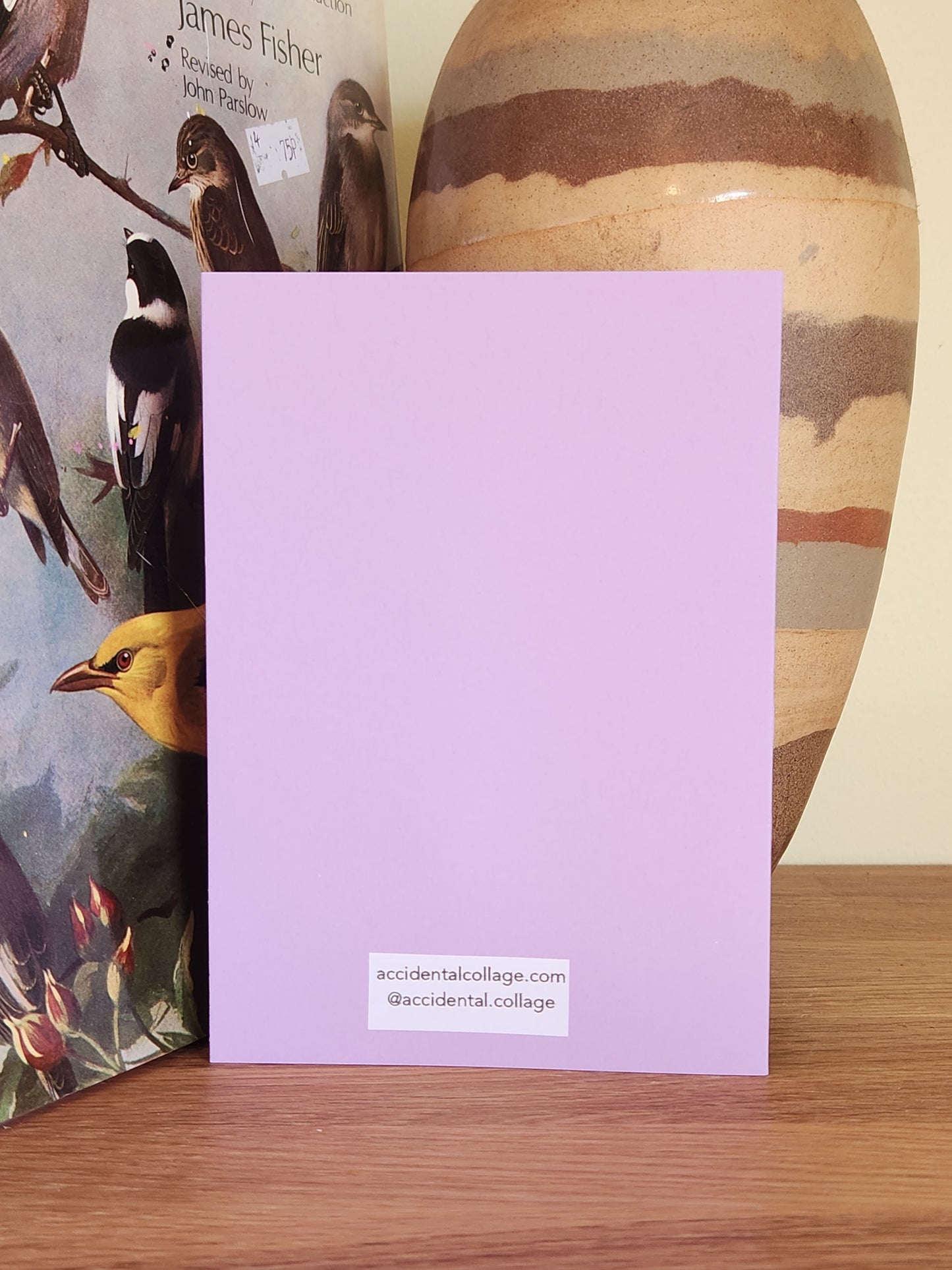 The back of a lavender greeting card standing upright on a wooden surface, with a small white label at the bottom reading 'accidentalkollage.com @accidental.collage'.