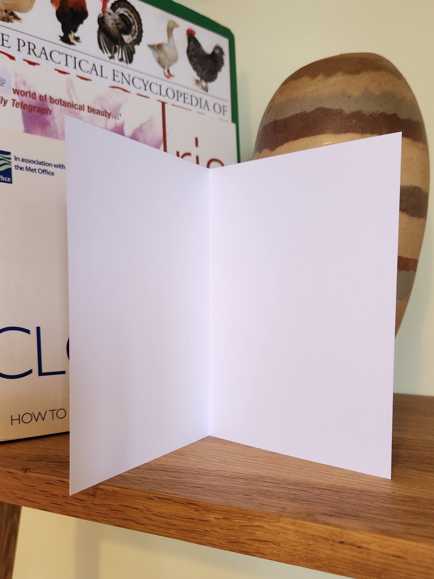 A blank white folded greeting card standing open on top of a wooden shelf next to several books.