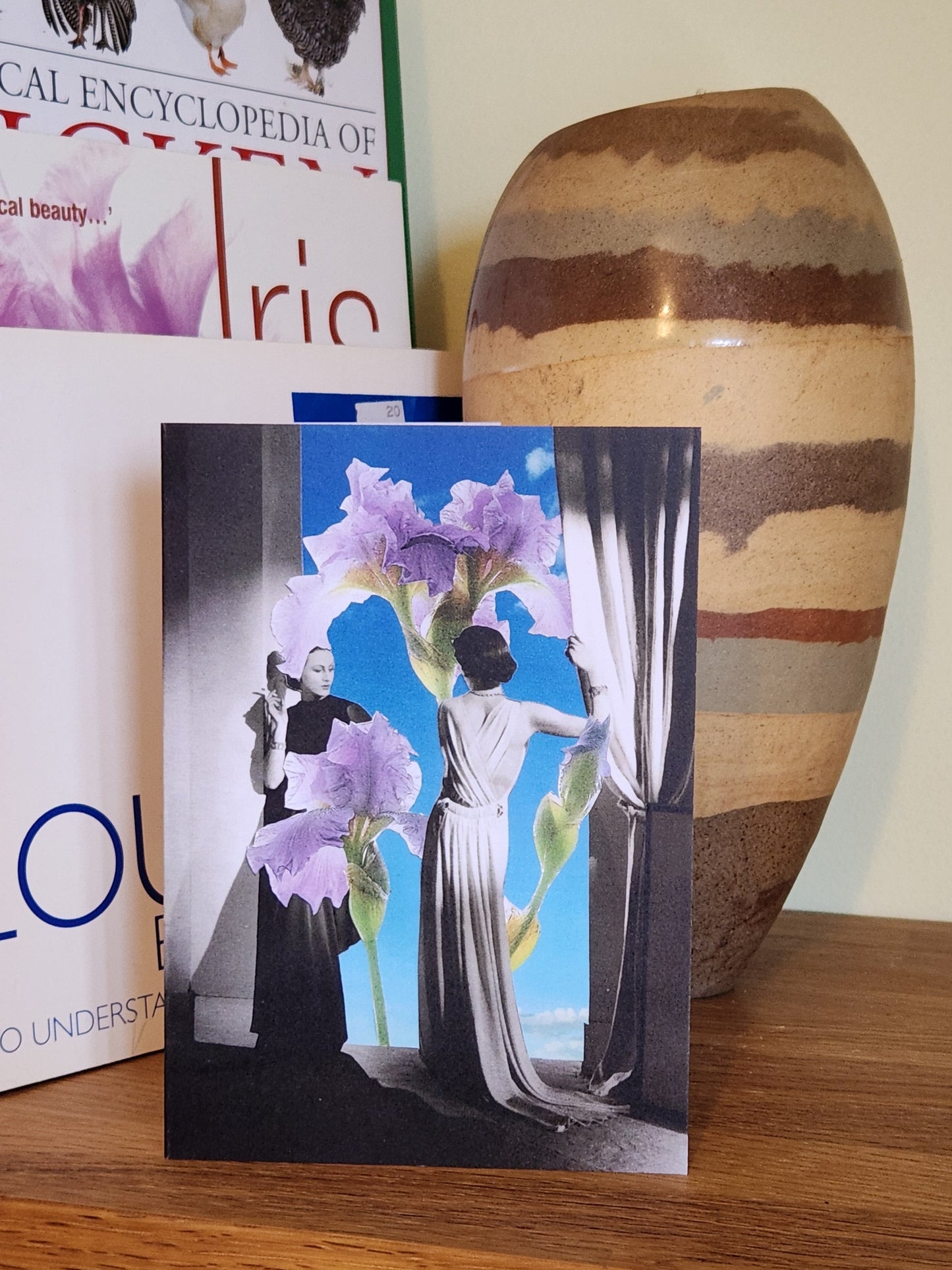 A greeting card featuring a collage print showing two women in long formal dresses standing in a room with curtains, facing a giant iris flower that reaches from the floor to the ceiling against a bright blue sky background, placed on a wooden shelf next to books and a striped ceramic vase.