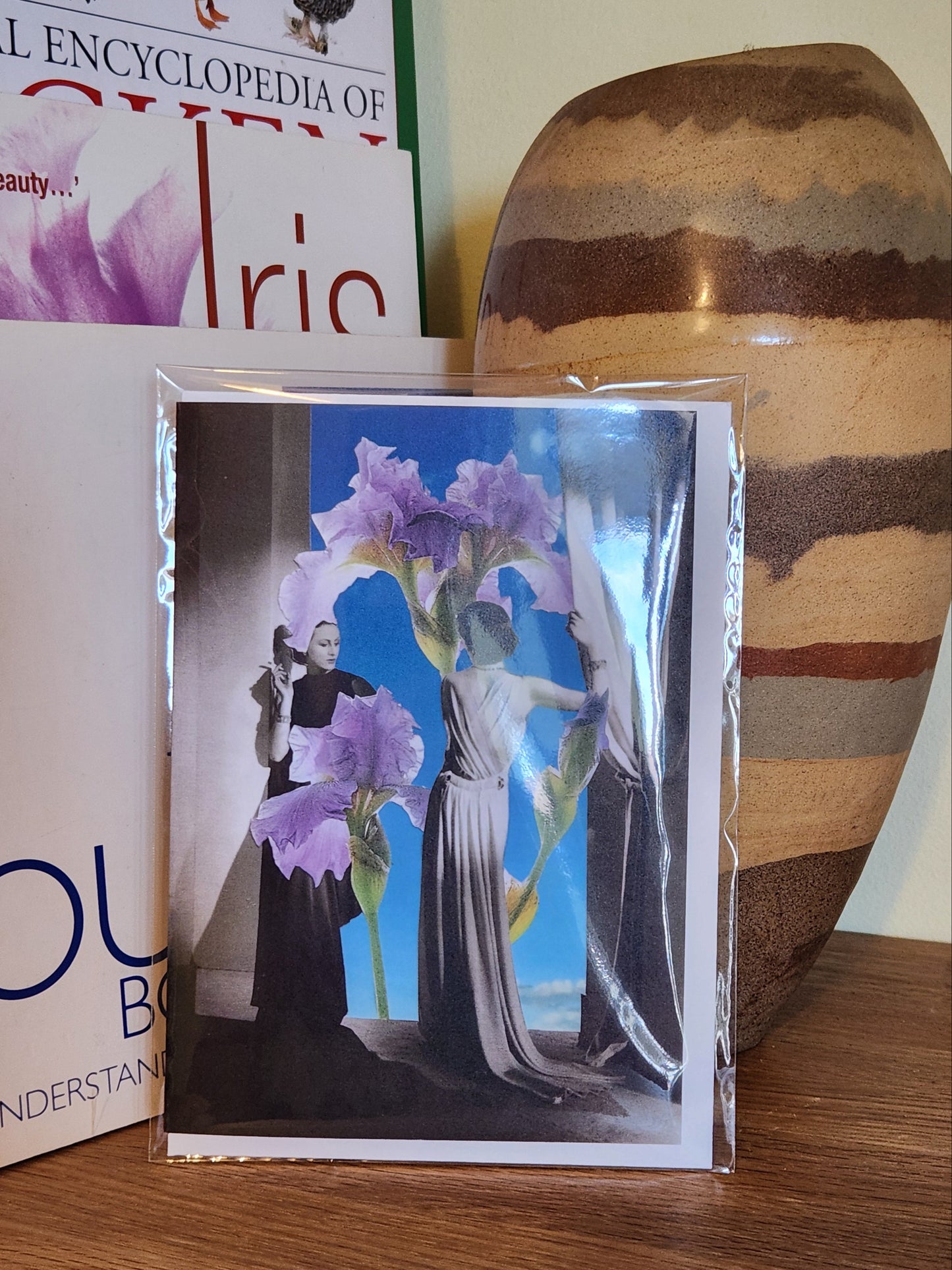 A greeting card featuring a collage print showing two women in long formal dresses standing in a room with curtains, facing a giant iris flower that reaches from the floor to the ceiling against a bright blue sky background, placed on a wooden shelf next to books and a striped ceramic vase, sealed in a clear plastic sleeve. 