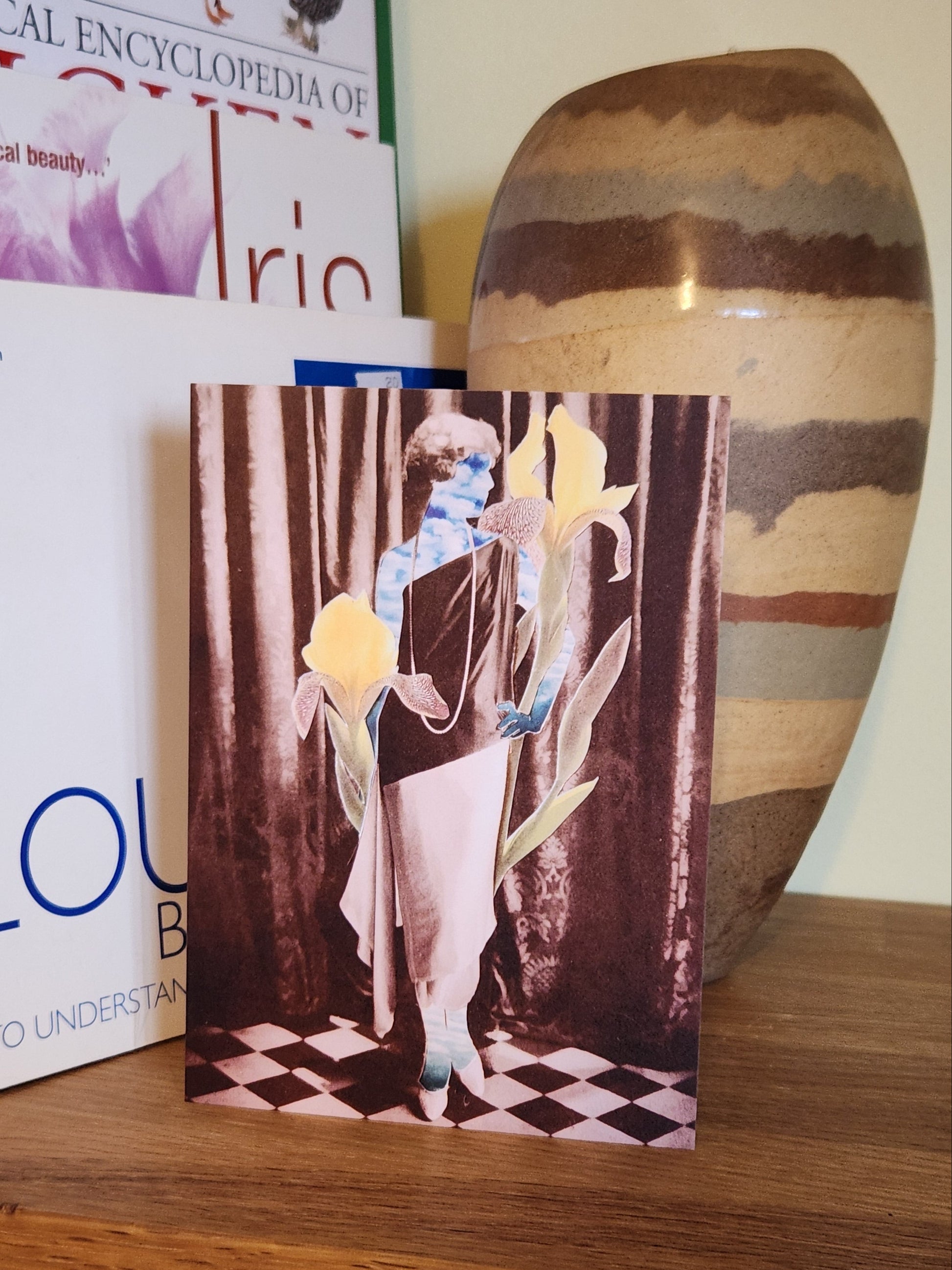A greeting card featuring a collage print of a woman made of sky standing in front of a dark curtain, wearing a long draped dress in black and white, with oversized yellow irises and green leaves around her, placed on a wooden shelf next to books and a striped ceramic vase.