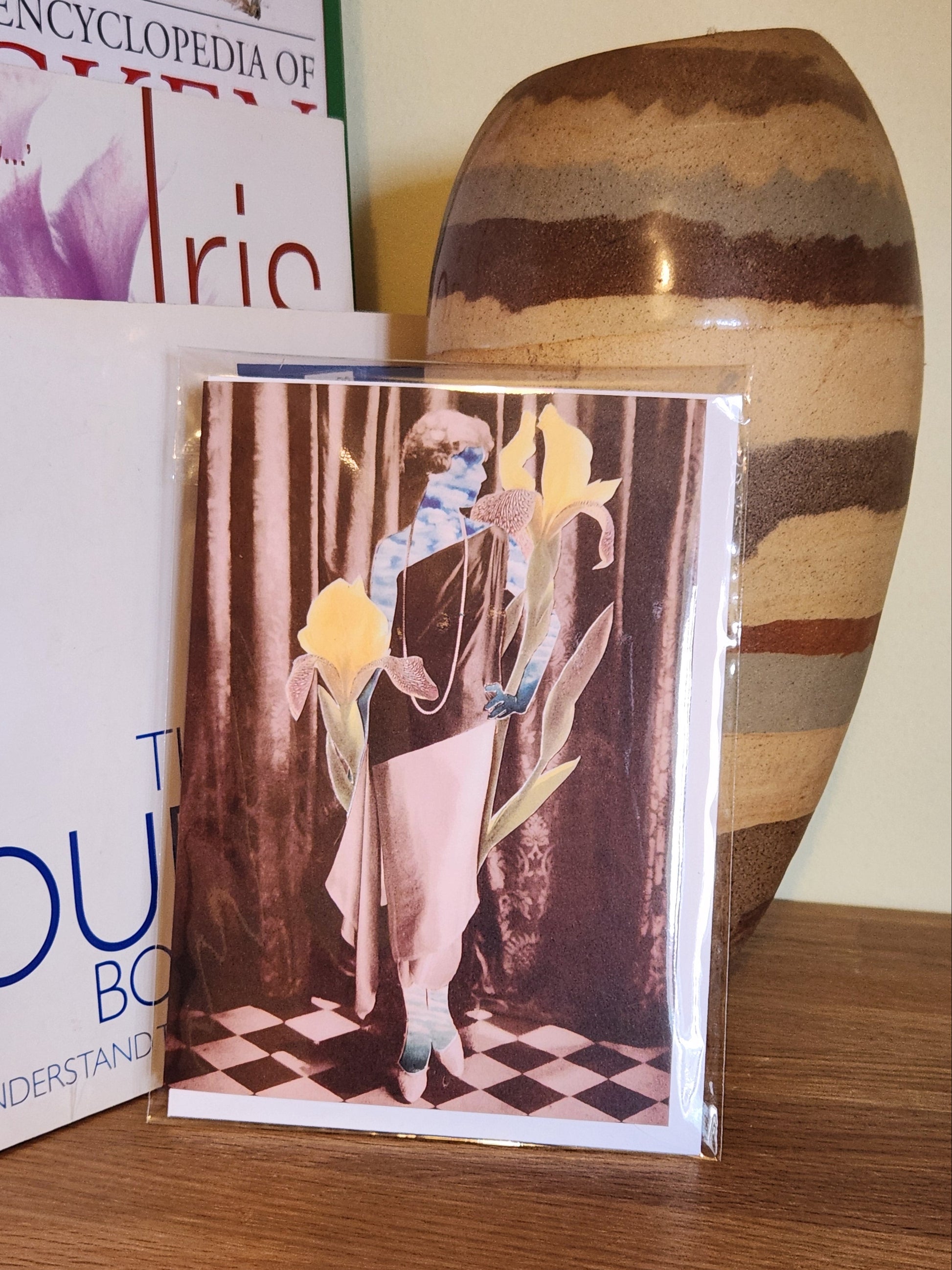 A greeting card featuring a collage print of a woman made of sky standing in front of a dark curtain, wearing a long draped dress in black and white, with oversized yellow irises and green leaves around her, placed on a wooden shelf next to books and a striped ceramic vase, sealed in a clear plastic sleeve.