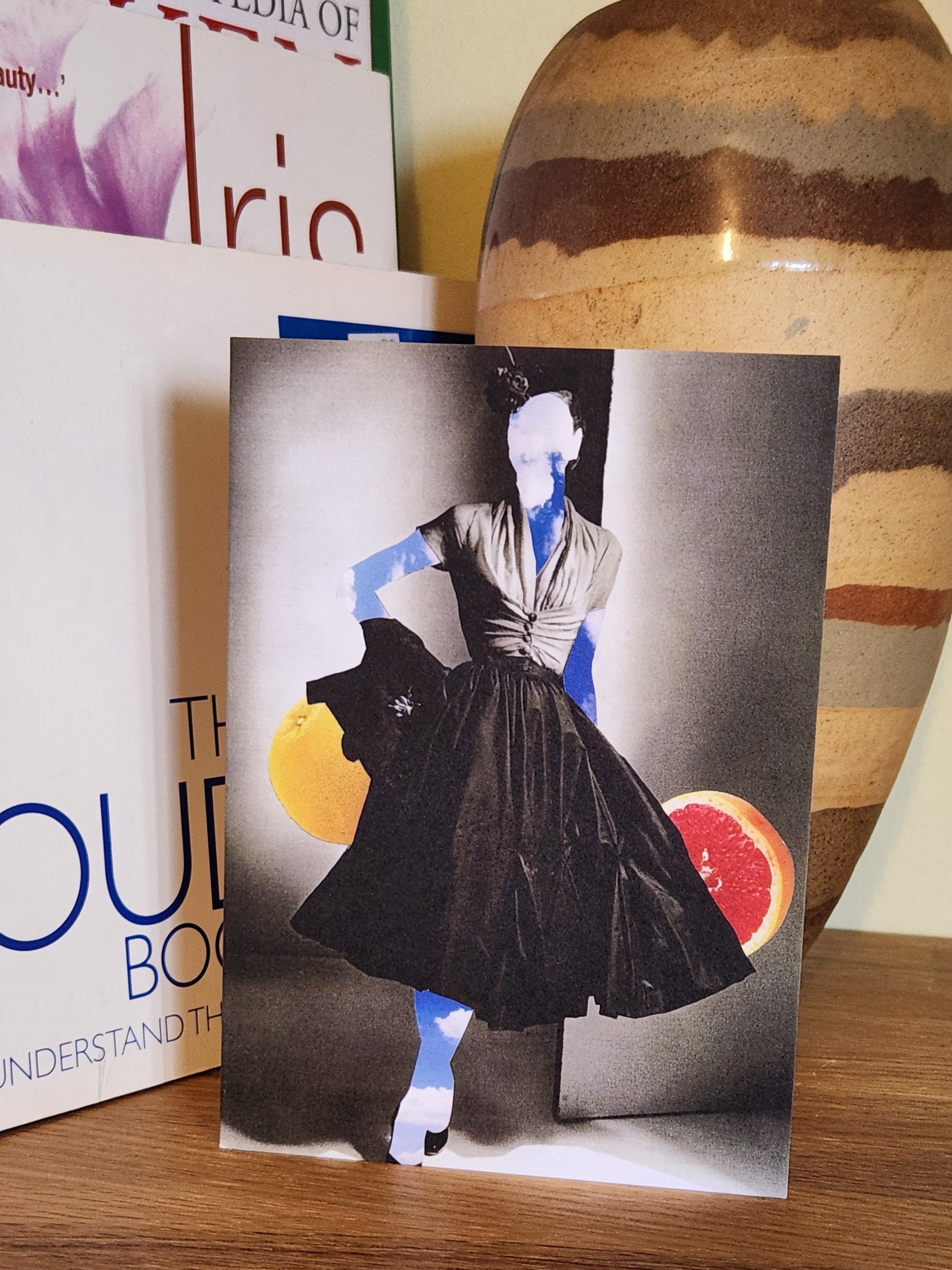 A greeting card featuring a collage print of a woman made of sky wearing a black pleated dress, with a gray background and fruit slices behind her, placed on a wooden shelf next to books and a striped ceramic vase.