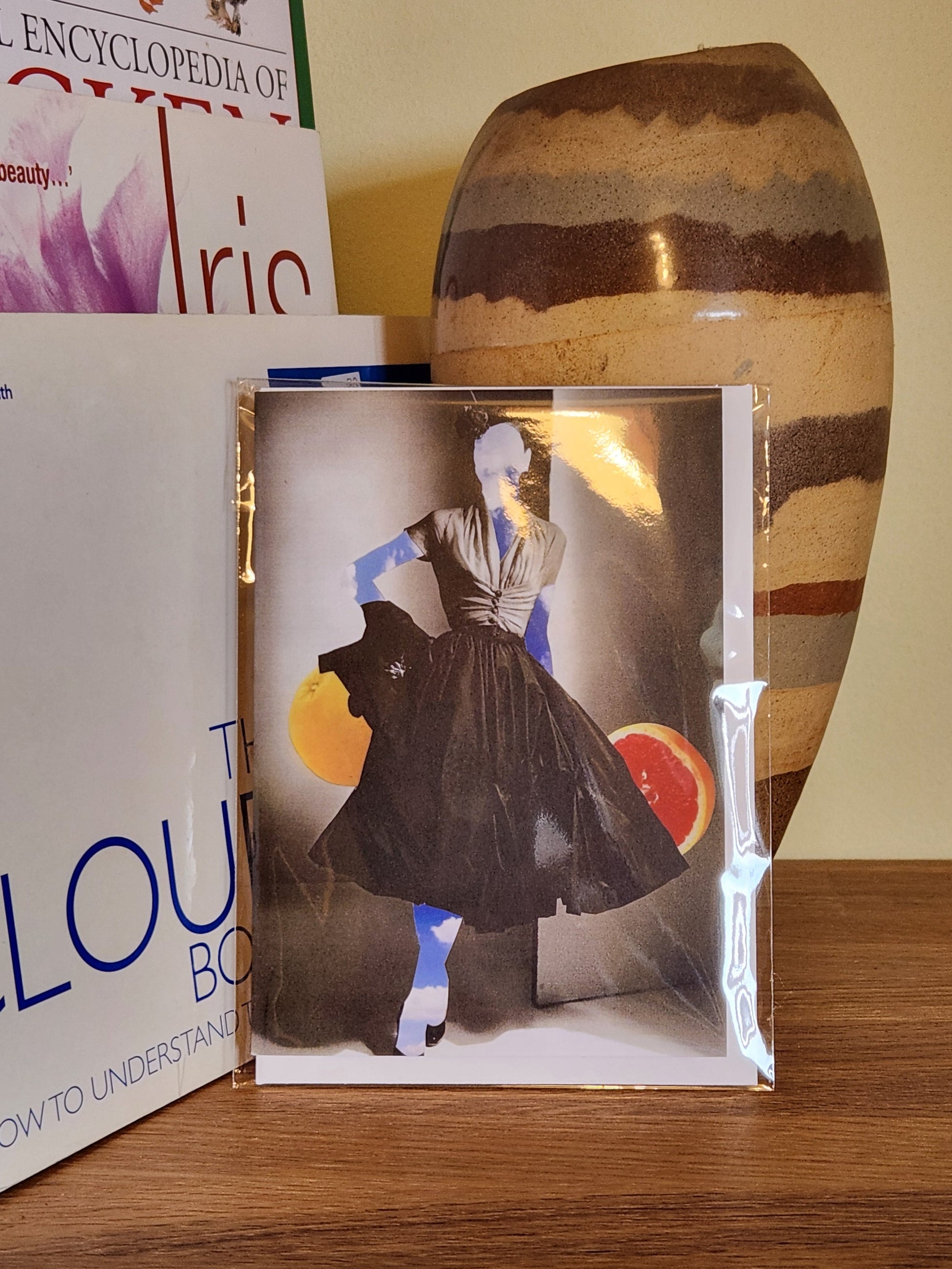 A greeting card featuring a collage print of a woman made of sky wearing a black pleated dress, with a gray background and fruit slices behind her, placed on a wooden shelf next to books and a striped ceramic vase, sealed in a clear plastic sleeve