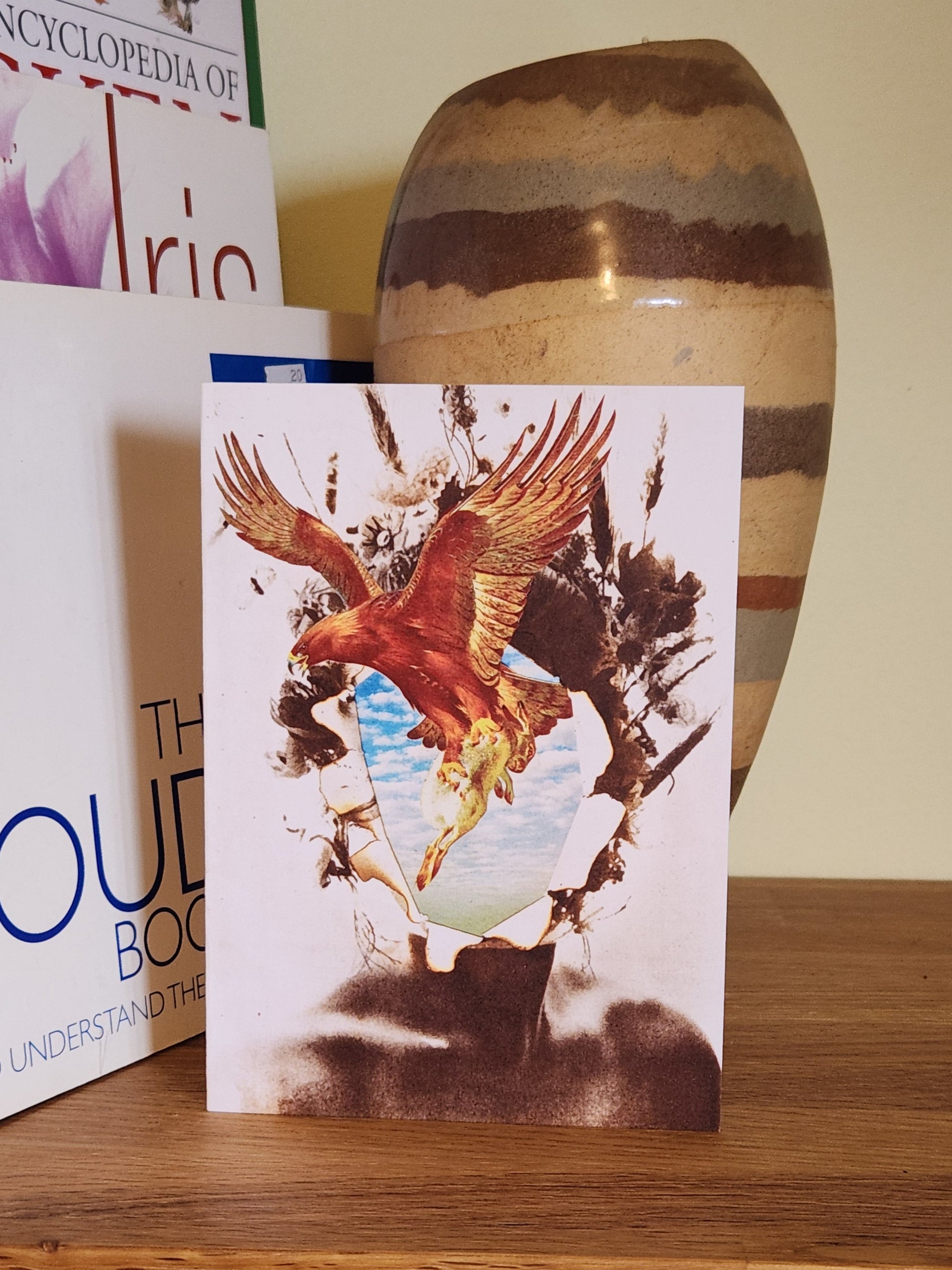 A greeting card featuring a collage print of a bird in flight, placed on a wooden shelf next to books and a striped ceramic vase.