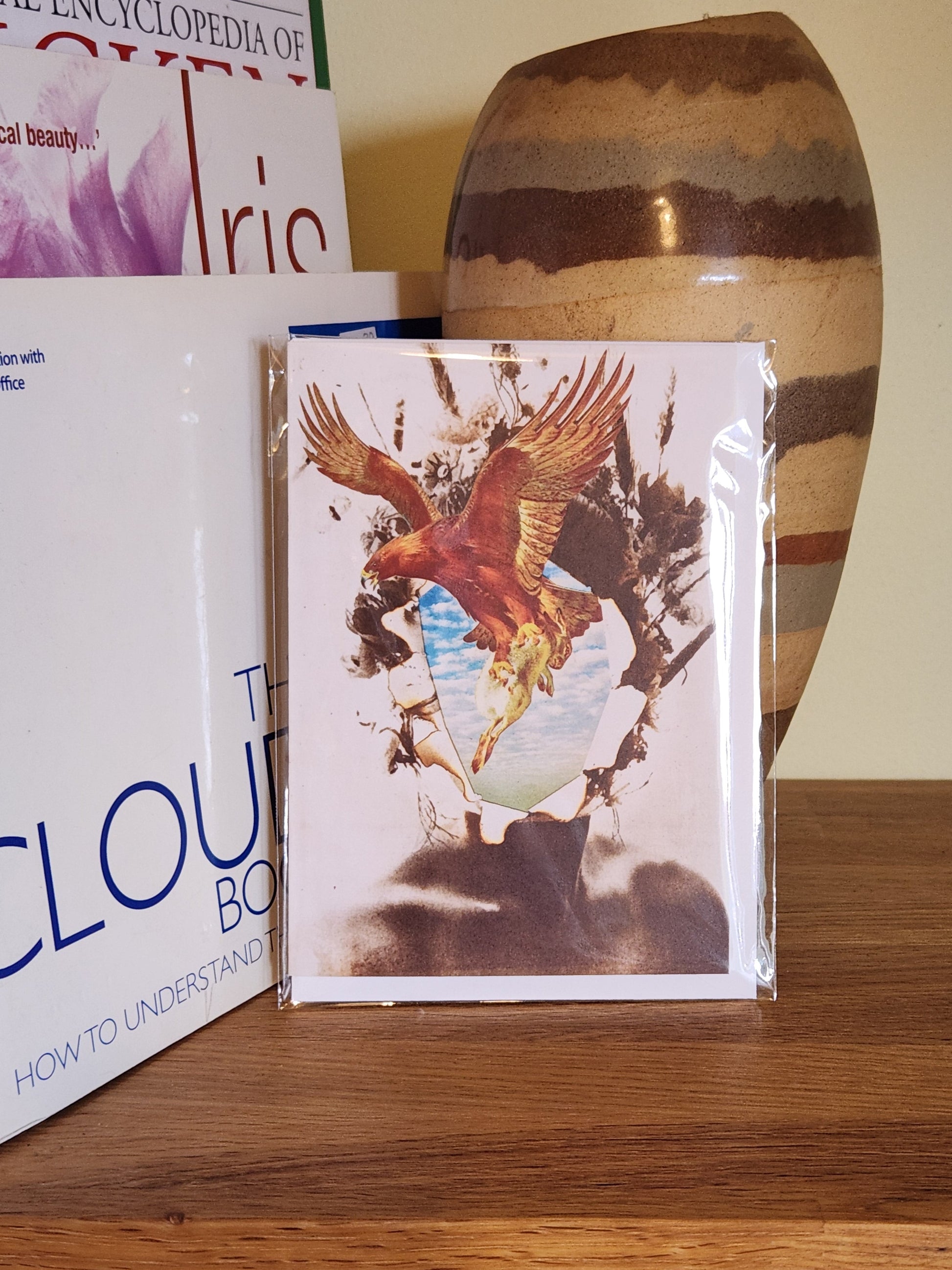 A greeting card featuring a collage print of a bird in flight, placed on a wooden shelf next to books and a striped ceramic vase, sealed in a clear plastic sleeve.