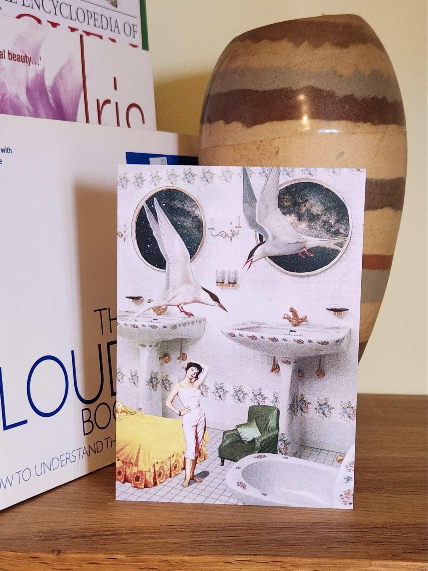 A greeting card featuring a collage print of terns, Natalie Wood and bedroom furniture in a white bathroom, placed on a wooden shelf next to books and a striped ceramic vase.