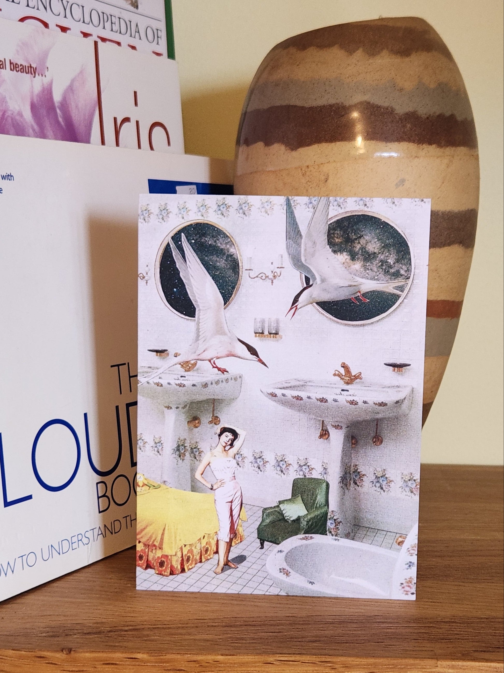 A greeting card featuring a collage print of terns, Natalie Wood and bedroom furniture in a white bathroom, placed on a wooden shelf next to books and a striped ceramic vase.