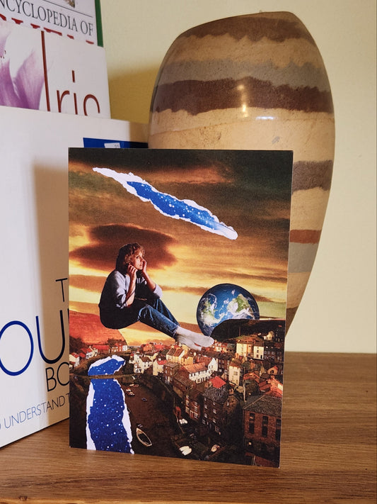 A greeting card featuring a surreal collage print showing a seated person floating above a coastal town, with a large Earth visible in the sky, dramatic sunset clouds, and torn blue-and-white paper effects integrated into the scene, placed on a wooden shelf next to books and a striped ceramic vase.