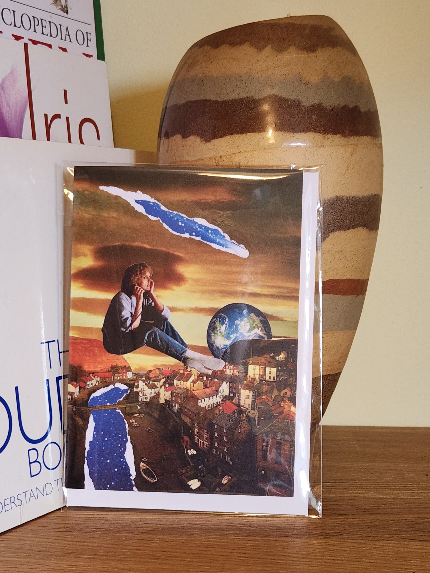 A greeting card featuring a surreal collage print showing a seated person floating above a coastal town, with a large Earth visible in the sky, dramatic sunset clouds, and torn blue-and-white paper effects integrated into the scene, placed on a wooden shelf next to books and a striped ceramic vase, sealed in a clear plastic sleeve.