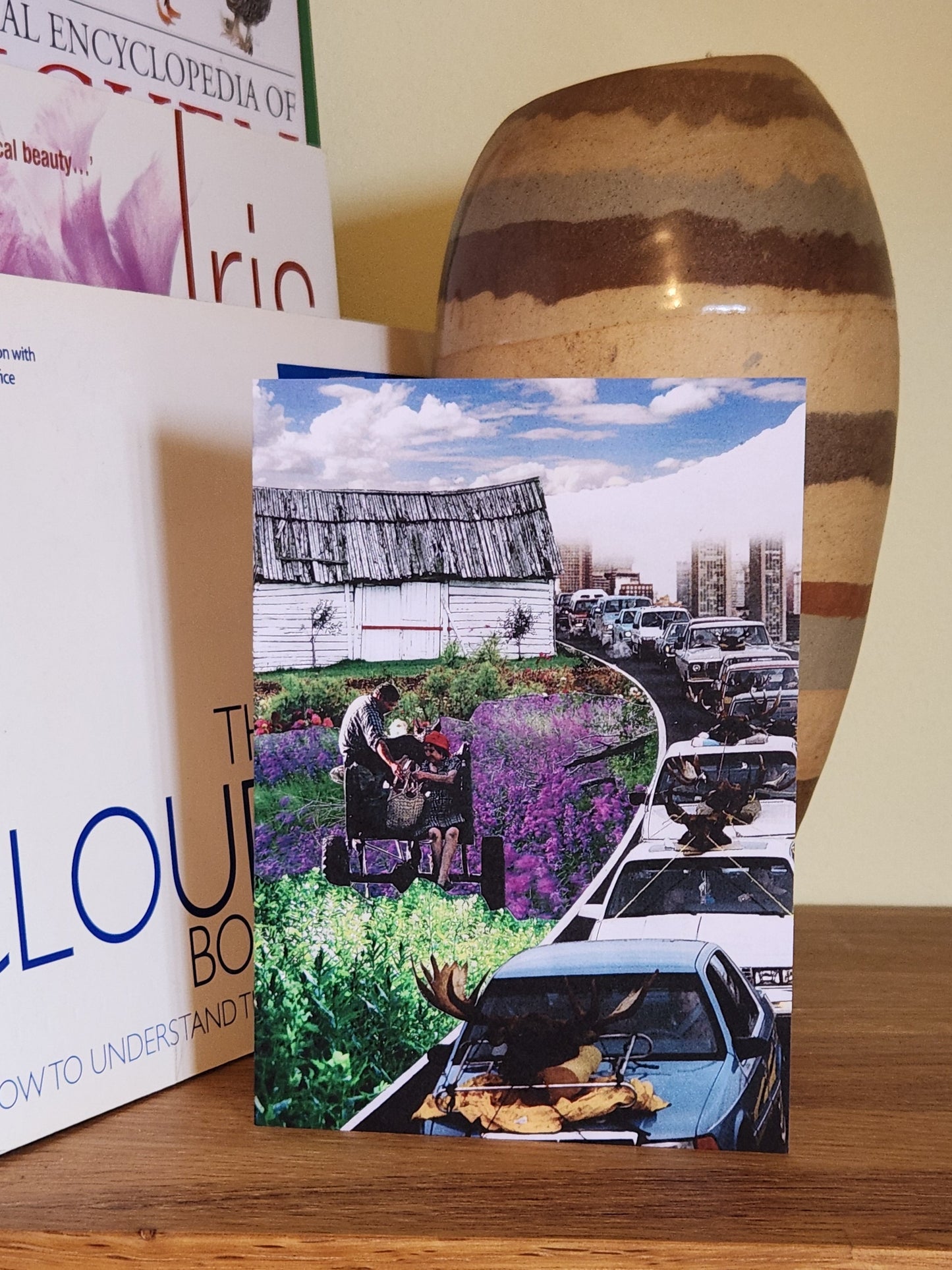 A greeting cards featuring a collage print of a surreal scene of a couple on a wagon in a field of purple flowers next to a highway with cars, displayed on a wooden shelf next to books and a ceramic vase.