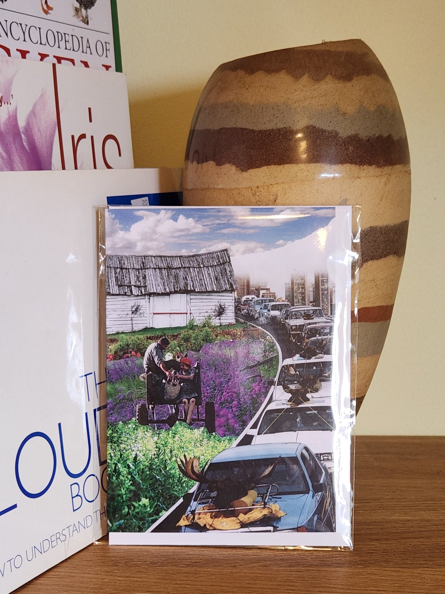 A greeting cards featuring a collage print of a surreal scene of a couple on a wagon in a field of purple flowers next to a highway with cars, displayed on a wooden shelf next to books and a ceramic vase, sealed in a clear plastic sleeve.