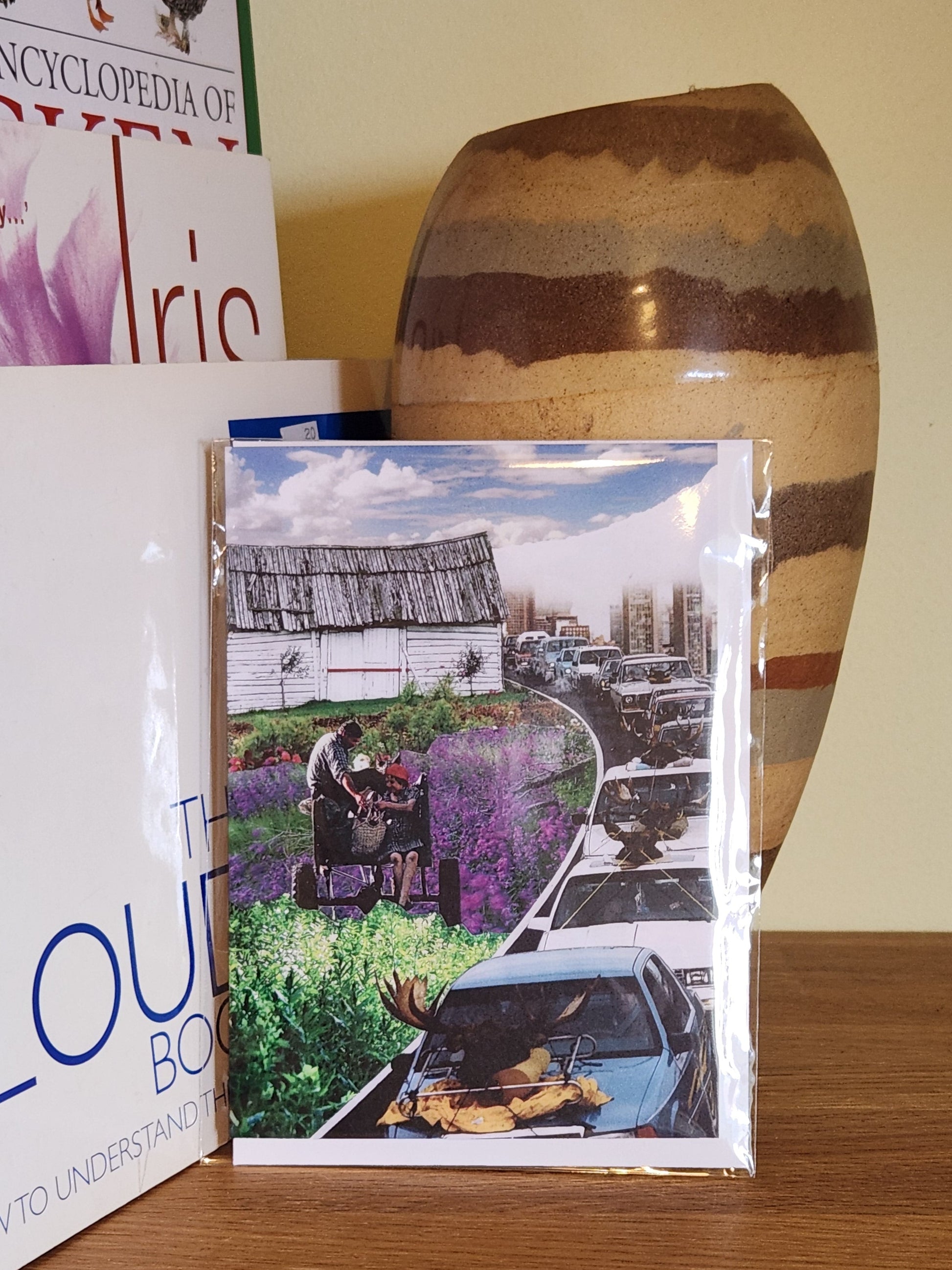 A greeting cards featuring a collage print of a surreal scene of a couple on a wagon in a field of purple flowers next to a highway with cars, displayed on a wooden shelf next to books and a ceramic vase, sealed in a clear plastic sleeve.