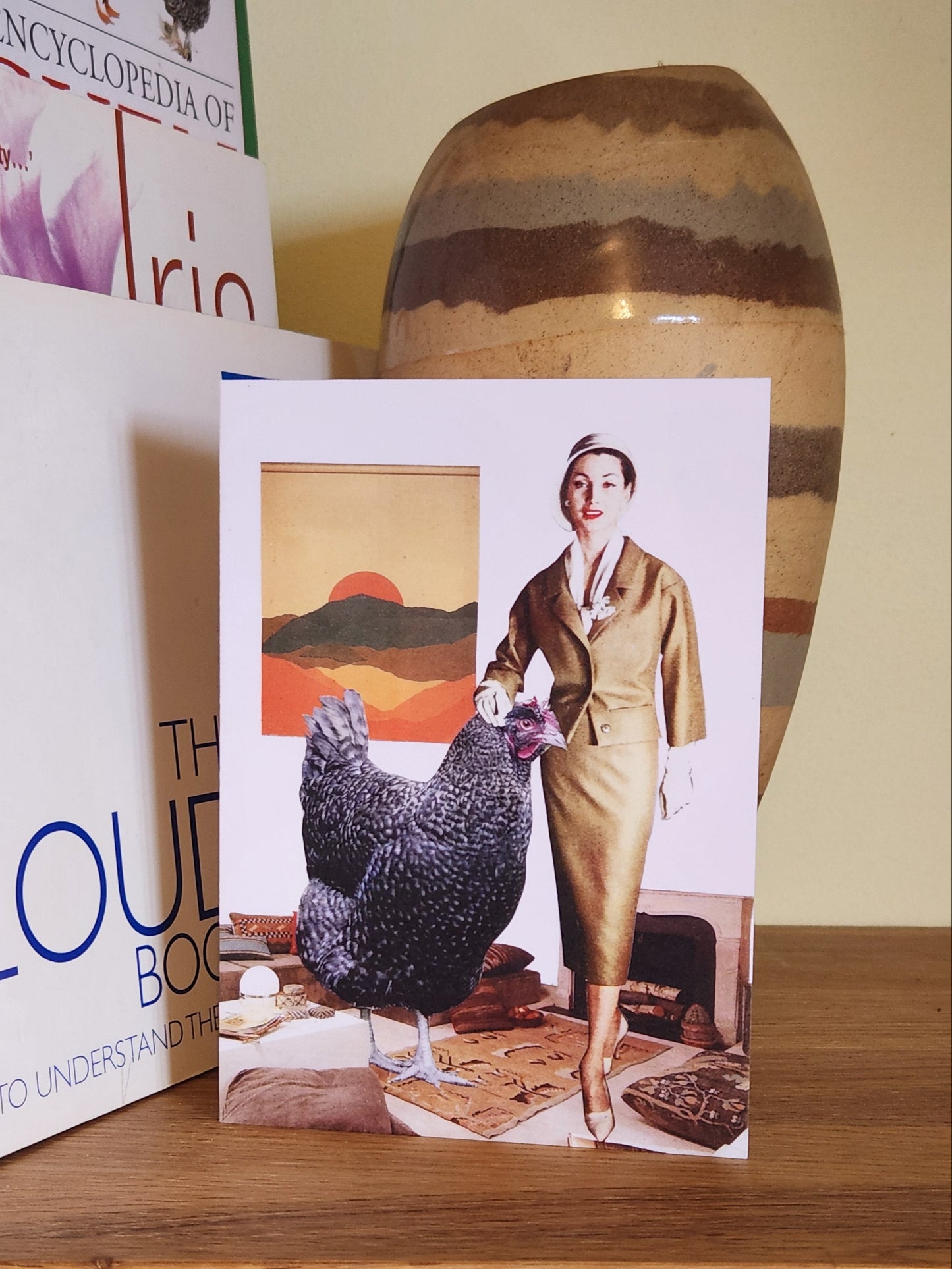 A greeting card featuring a collage print of a woman in a gold skirt suit stands indoors next to a very large black speckled chicken, with a framed mountain-and-sunset painting on the wall behind them, placed on a wooden shelf next to books and a striped ceramic vase.