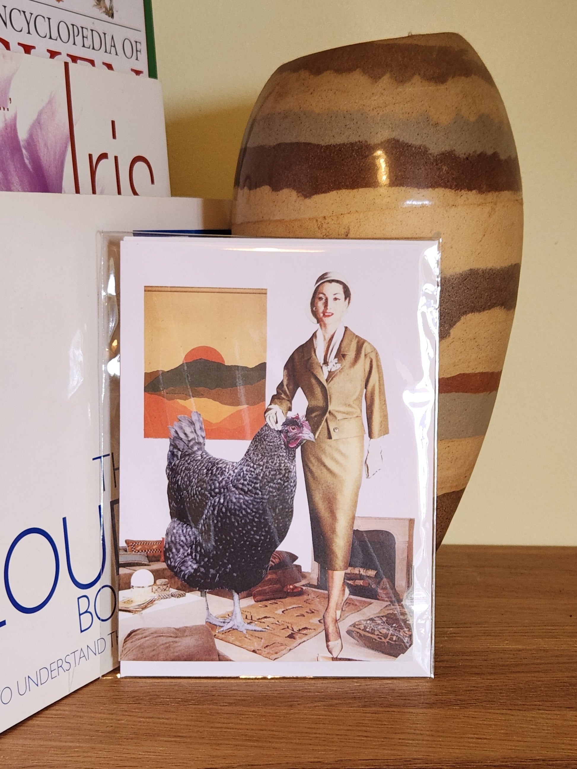 A greeting card featuring a collage print of a woman in a gold skirt suit stands indoors next to a very large black speckled chicken, with a framed mountain-and-sunset painting on the wall behind them, placed on a wooden shelf next to books and a striped ceramic vase, sealed in a clear plastic sleeve.