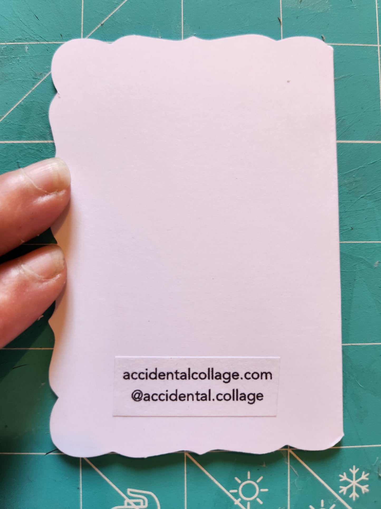 The back of a white card with scalloped edges against a green cutting mat. The card has a small vertical printed label on the bottomreading 'accidentalcollage.com, @accidental.collage'