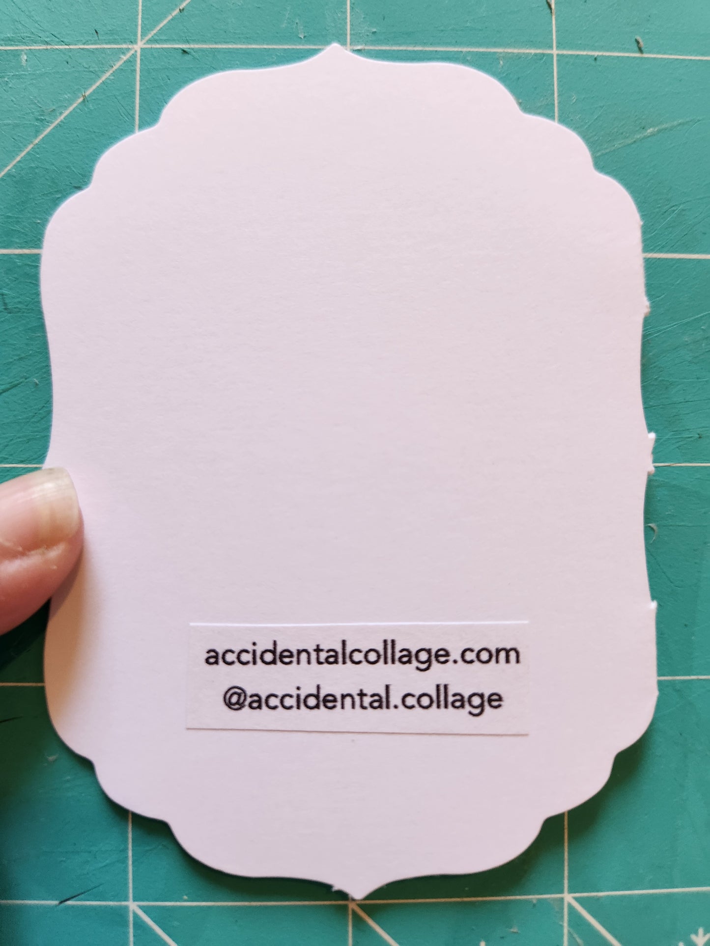 The back of a scalloped-edge white paper card with a small printed vertical text block reading 'accidentalcollage.com, @accidentalcollage' on a green cutting mat.