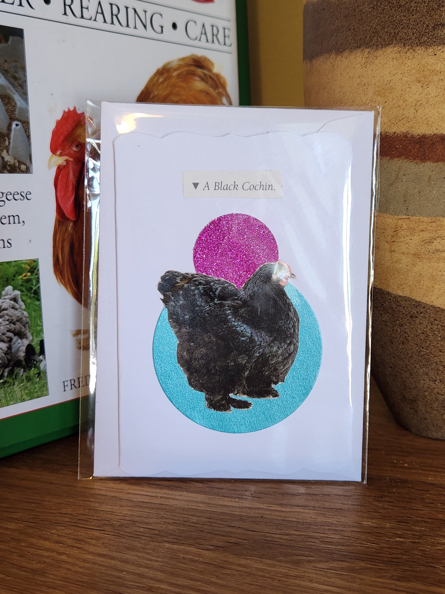 A printed card showing a black chicken over a pink and turquoise circle, in clear plastic packaging with matching envelope. The card is placed on a wooden shelf in front of a striped ceramic vase.