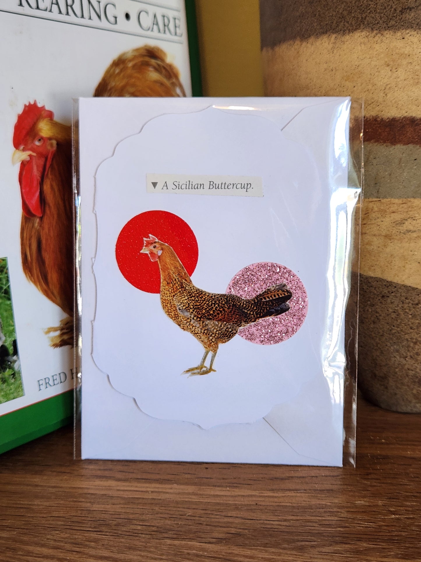 A scalloped white greeting card featuring a collage of a chicken over a red circle with a pink glitter circle behind it. The card is packaged in clear plastic with a matching envelope. The card is placed on a wooden shelf in front of a striped ceramic vase.