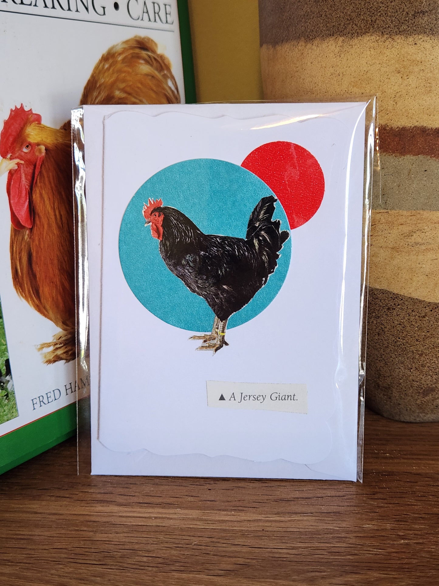 A decorative white greeting card featuring a black rooster over a turquoise and a red circle, in clear plastic packaging with matching envelope. The card is placed on a wooden shelf in front of a striped ceramic vase.