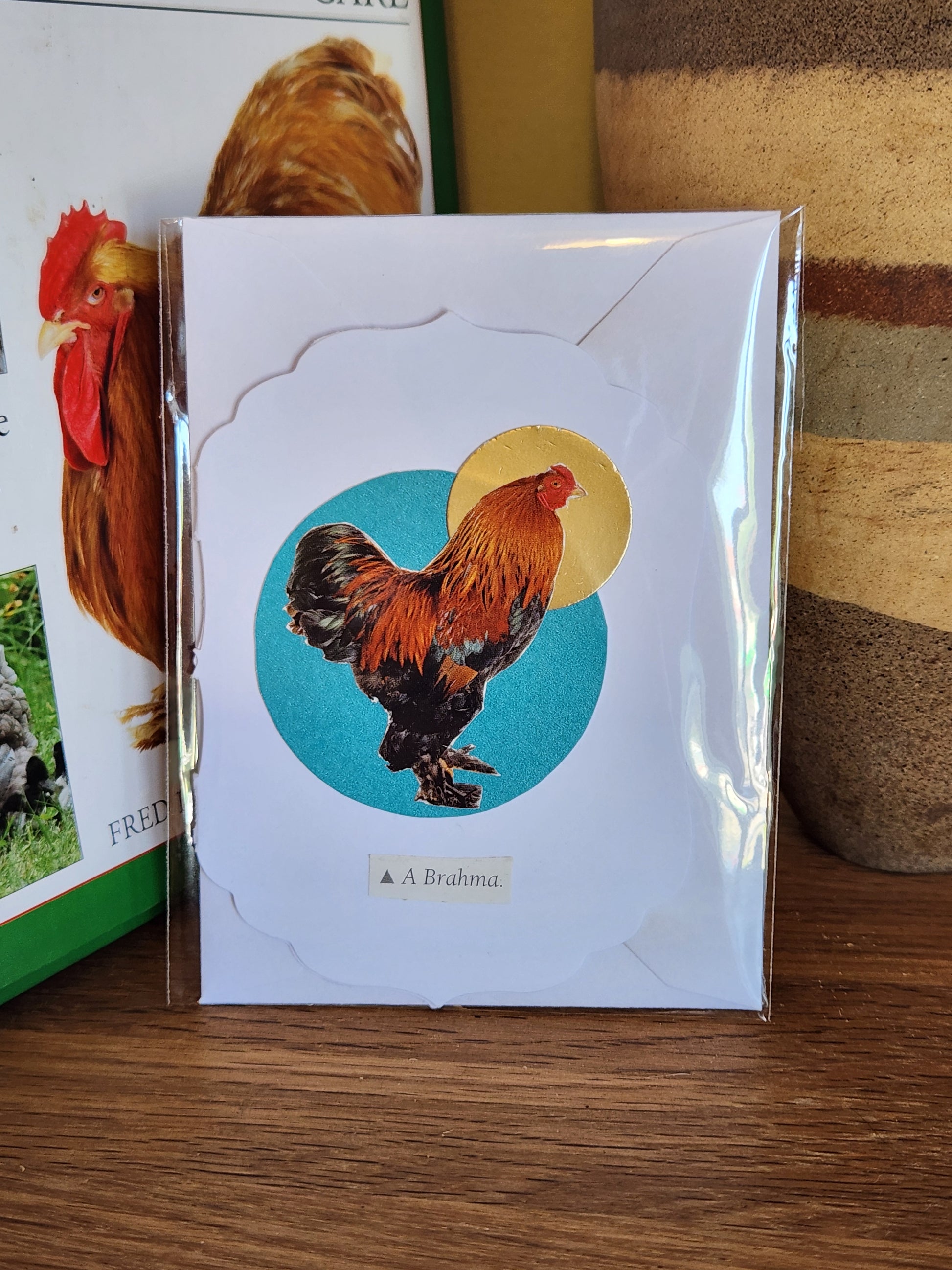 A small white art gretting card featuring a rooster collage over a turquoise and a gold circle, sealed in clear plastic packaging with matching envelope. The card is placed on a wooden shelf in front of a striped ceramic vase.