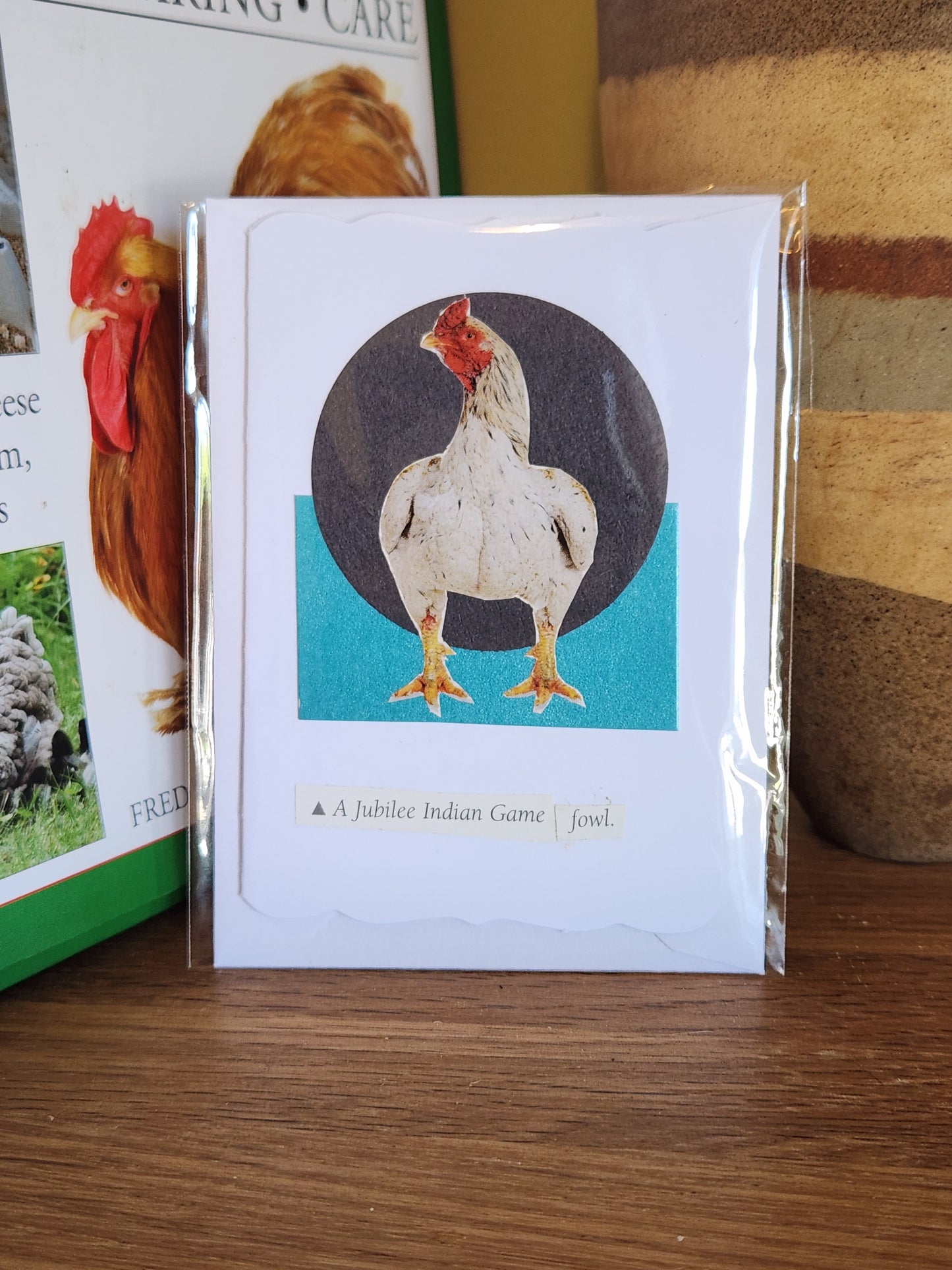 A small decorative card featuring a white chicken illustration over a dark circle and turquoise block, in clear plastic packaging with matching envelope. The card is placed on a wooden shelf in front of a striped ceramic vase.