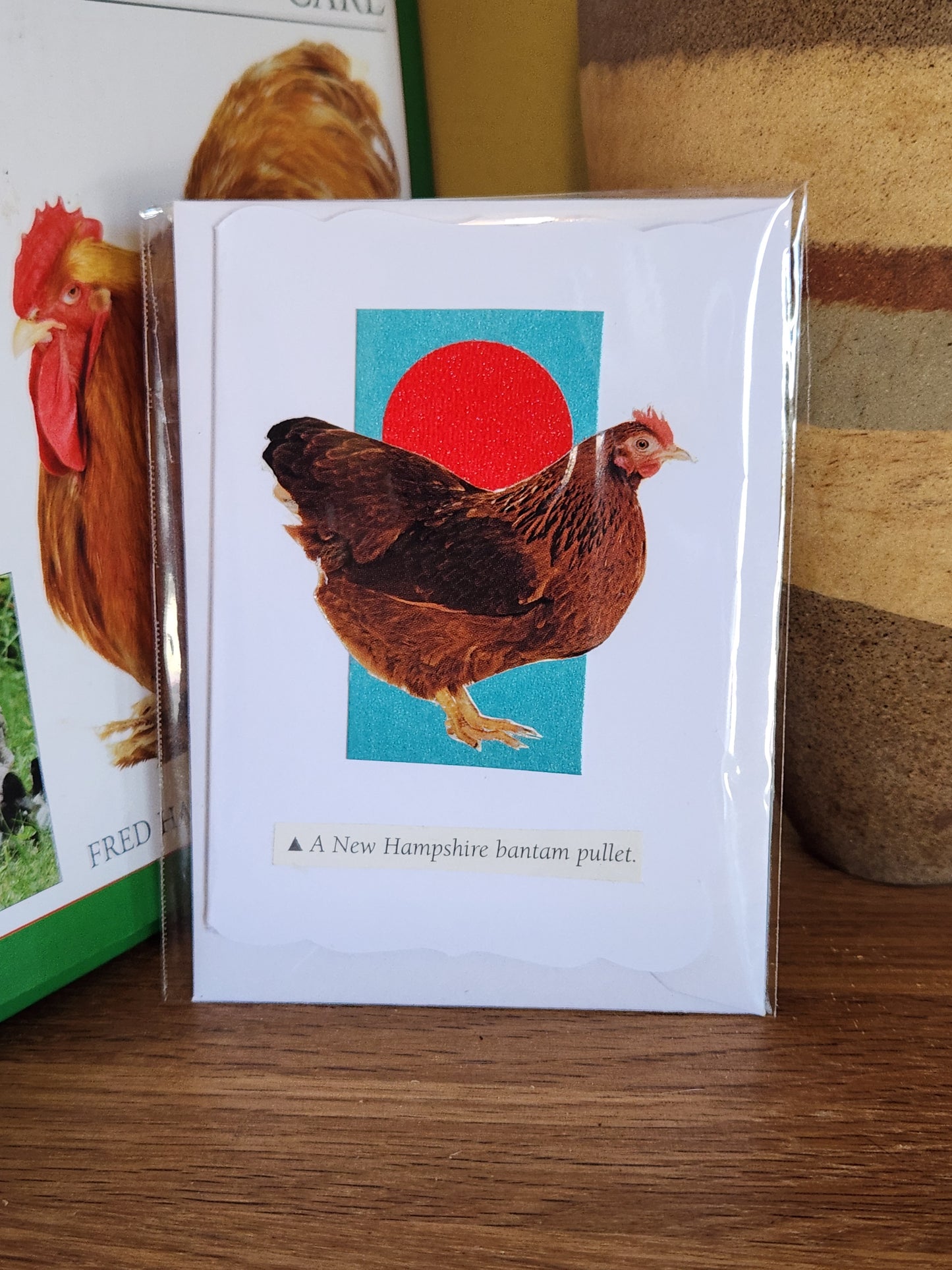 A card featuring a brown hen in front of a turquoise square with a red circle, in clear plastic packaging with matching envelope. The card is placed on a wooden shelf in front of a striped ceramic vase.