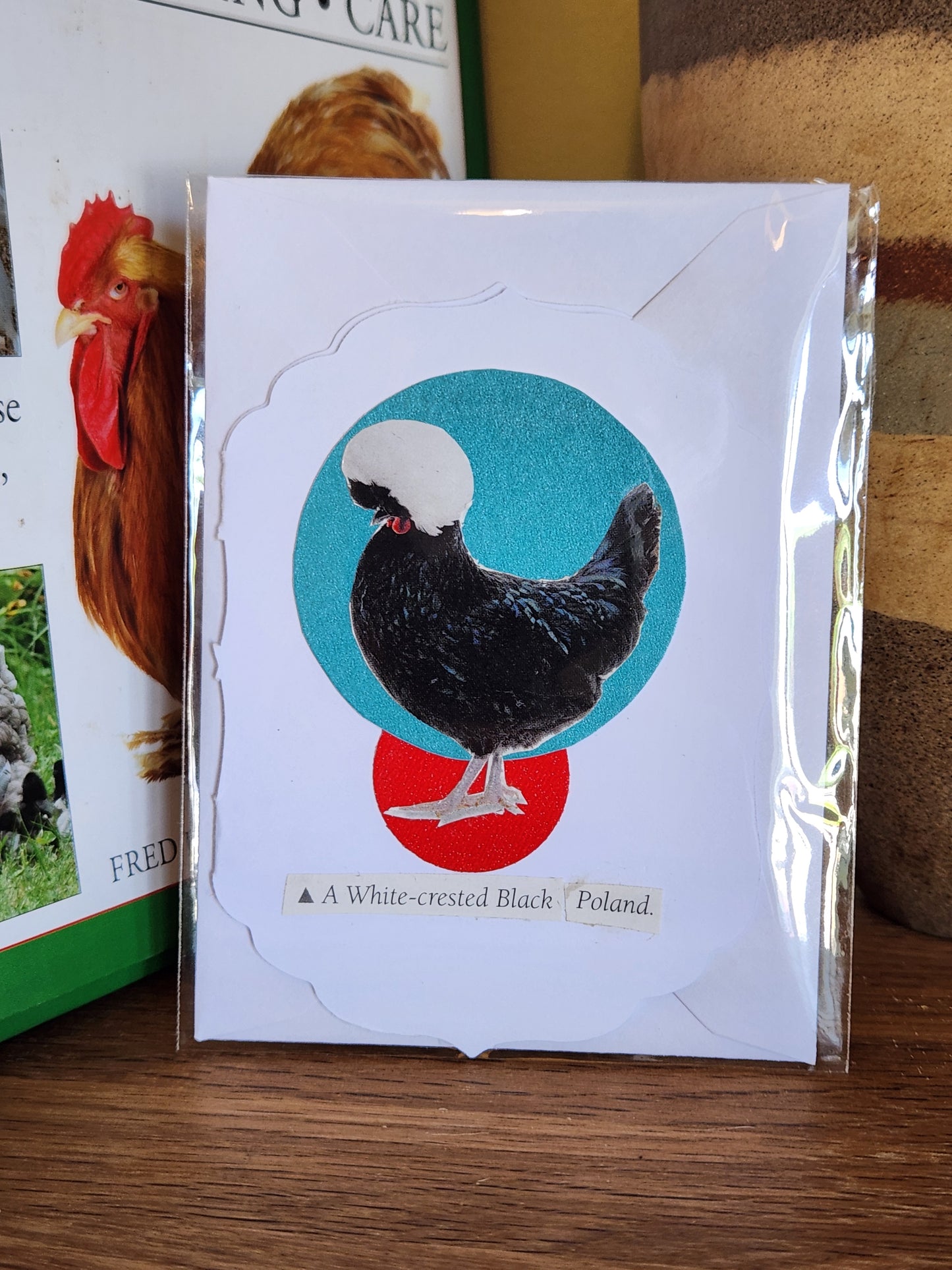 A small white card featuring a black-and-white rooster over a turquoise and red circular background, in clear plastic packaging with matching envelope. The card is placed on a wooden shelf in front of a striped ceramic vase.