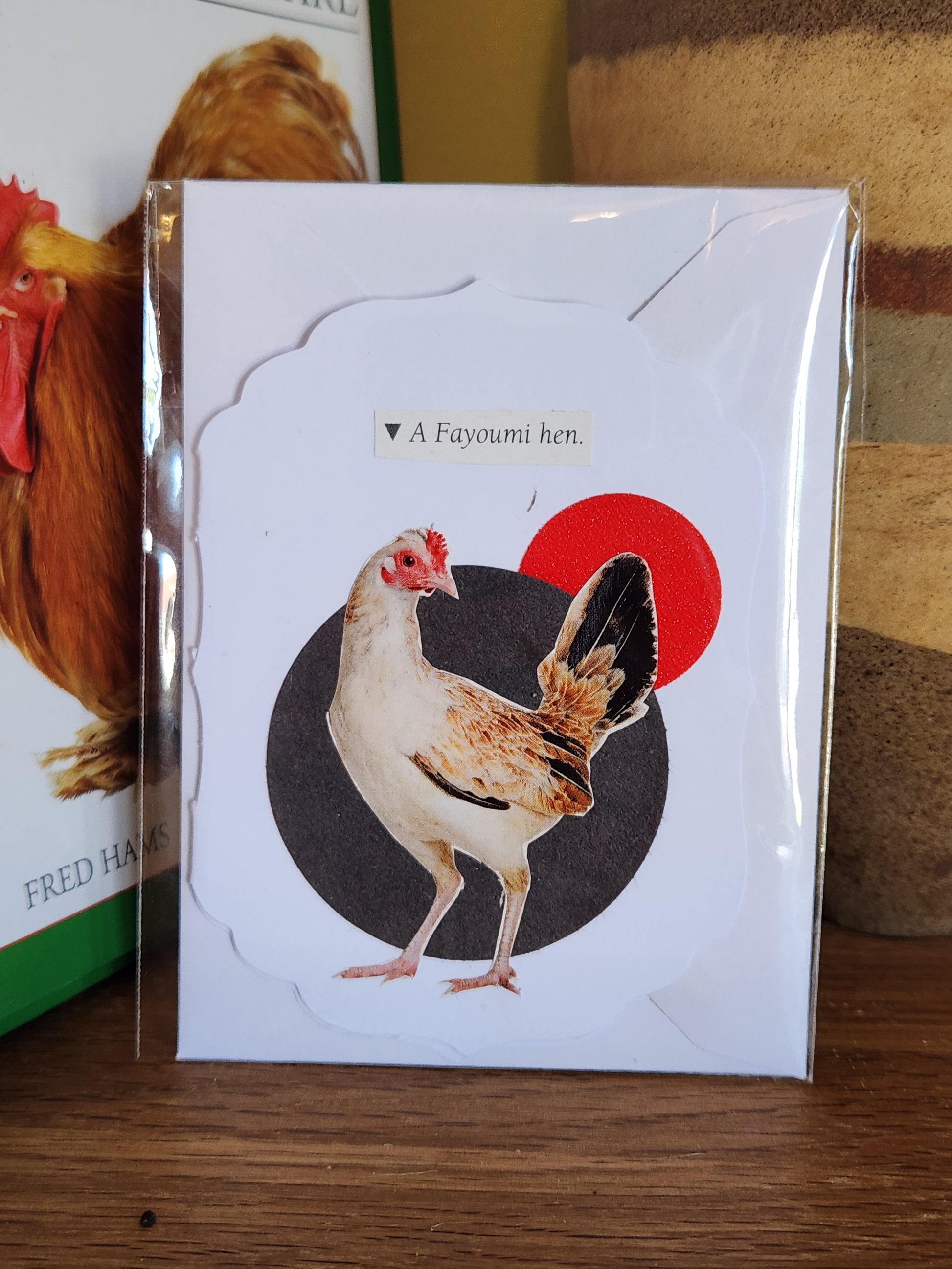 A small white greeting card featuring a collage of a chicken over a black and a red circle, in clear plastic packaging with matching envelope. The card is placed on a wooden shelf in front of a striped ceramic vase.