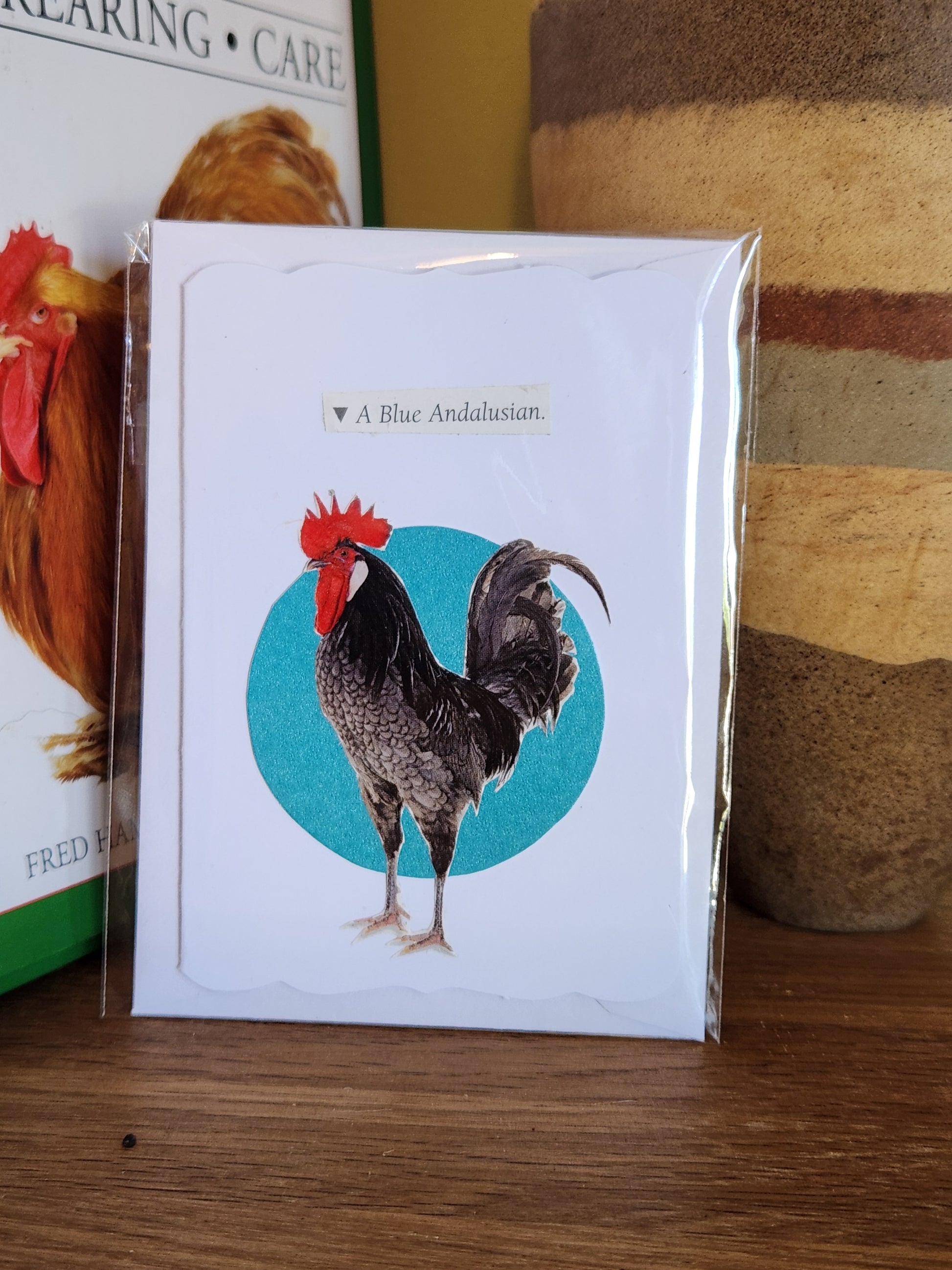A collage card featuring a rooster over a turquoise circle, in clear plastic packaging with matching envelope. The card is placed on a wooden shelf in front of a striped ceramic vase.