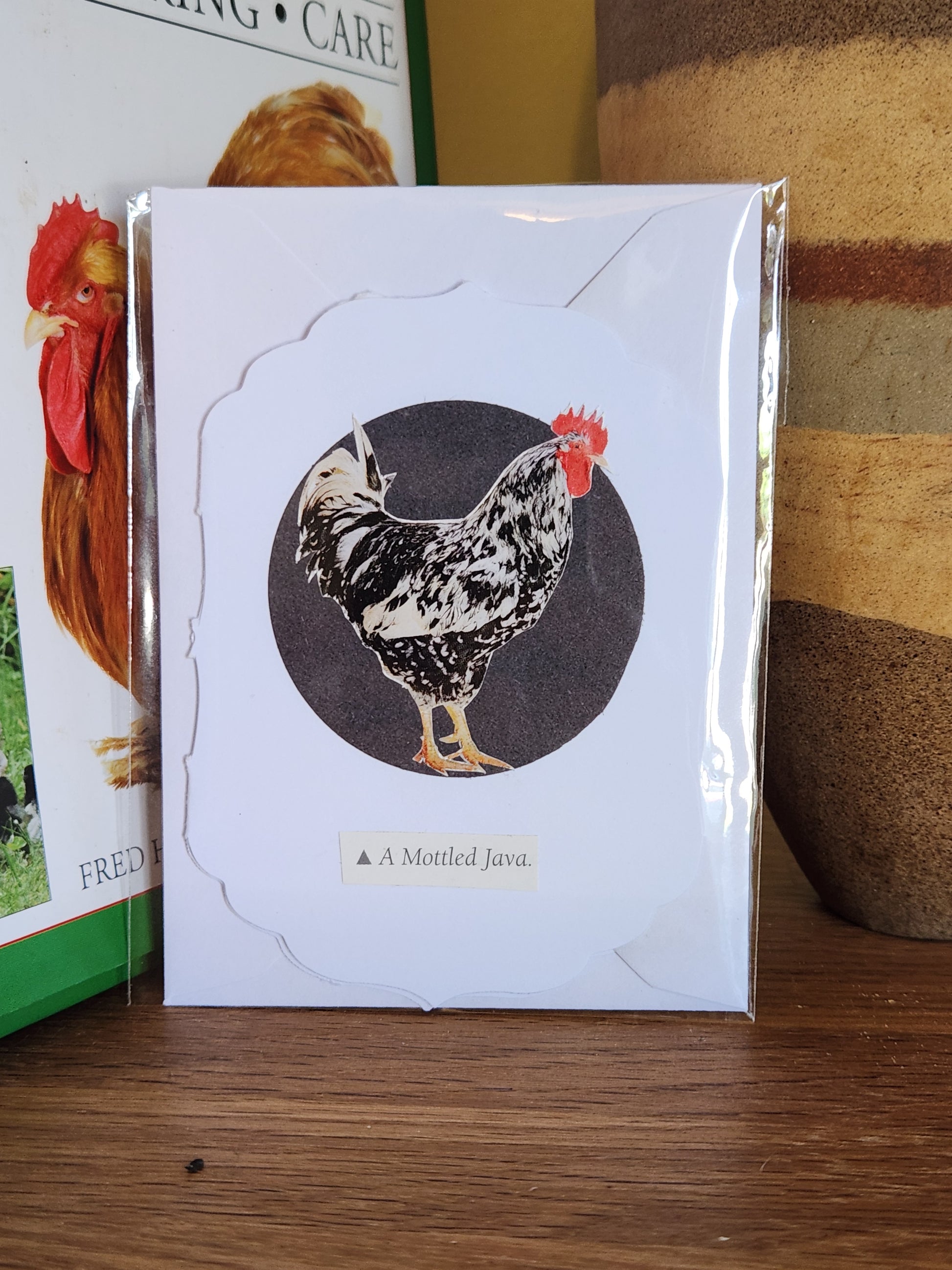 A small white greeting card featuring a black-and-white rooster on a black circle with the text 'A Mottle Java' underneath, in clear plastic packaging with matching envelope. The card is placed on a wooden shelf in front of a striped ceramic vase.