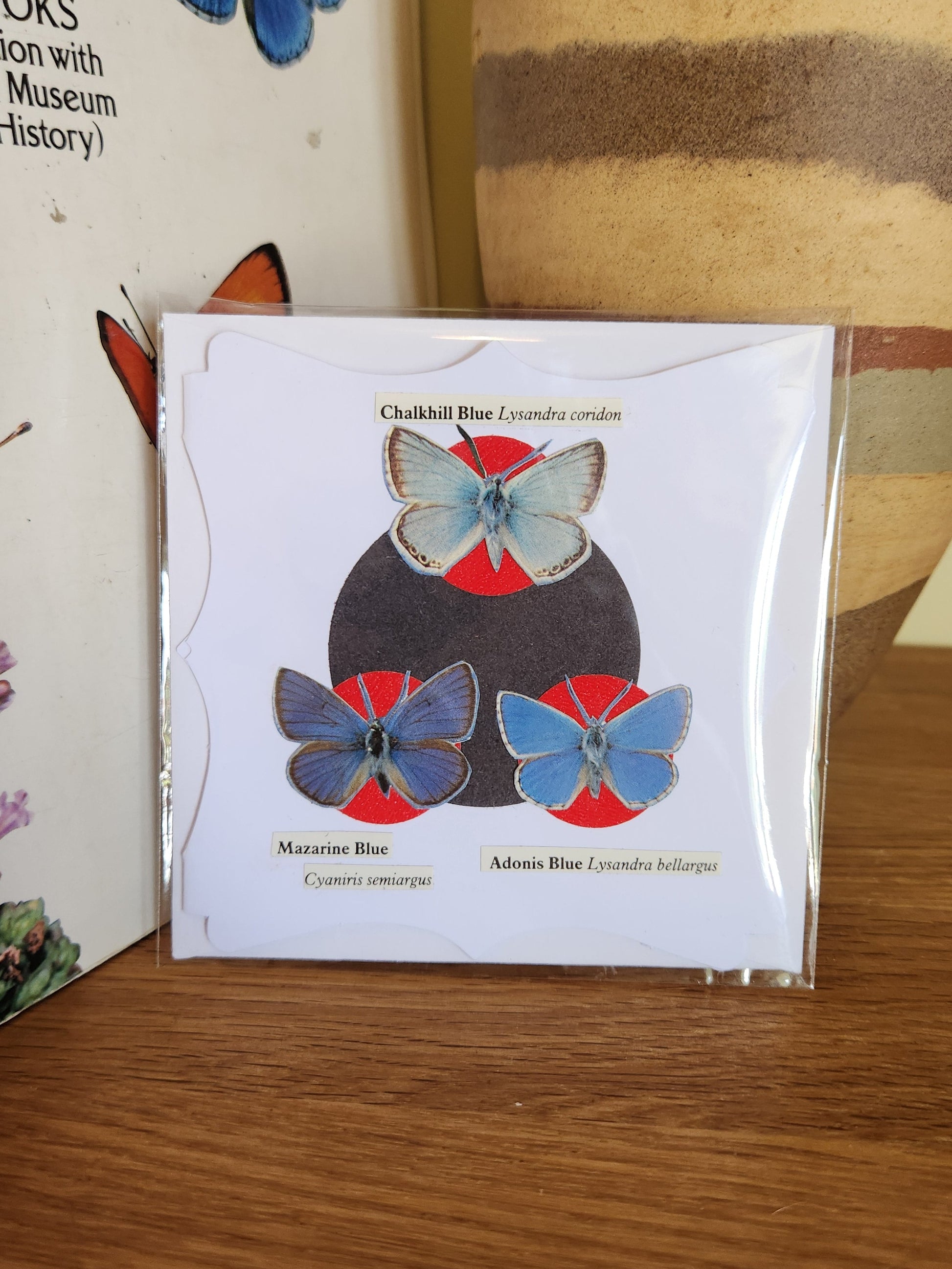 A decorative white greeting card featuring three blue butterflies over a black and a red circle motif, sealed in clear plastic packaging with matching envelope. The card is placed on a wooden shelf in front of a striped ceramic vase.
