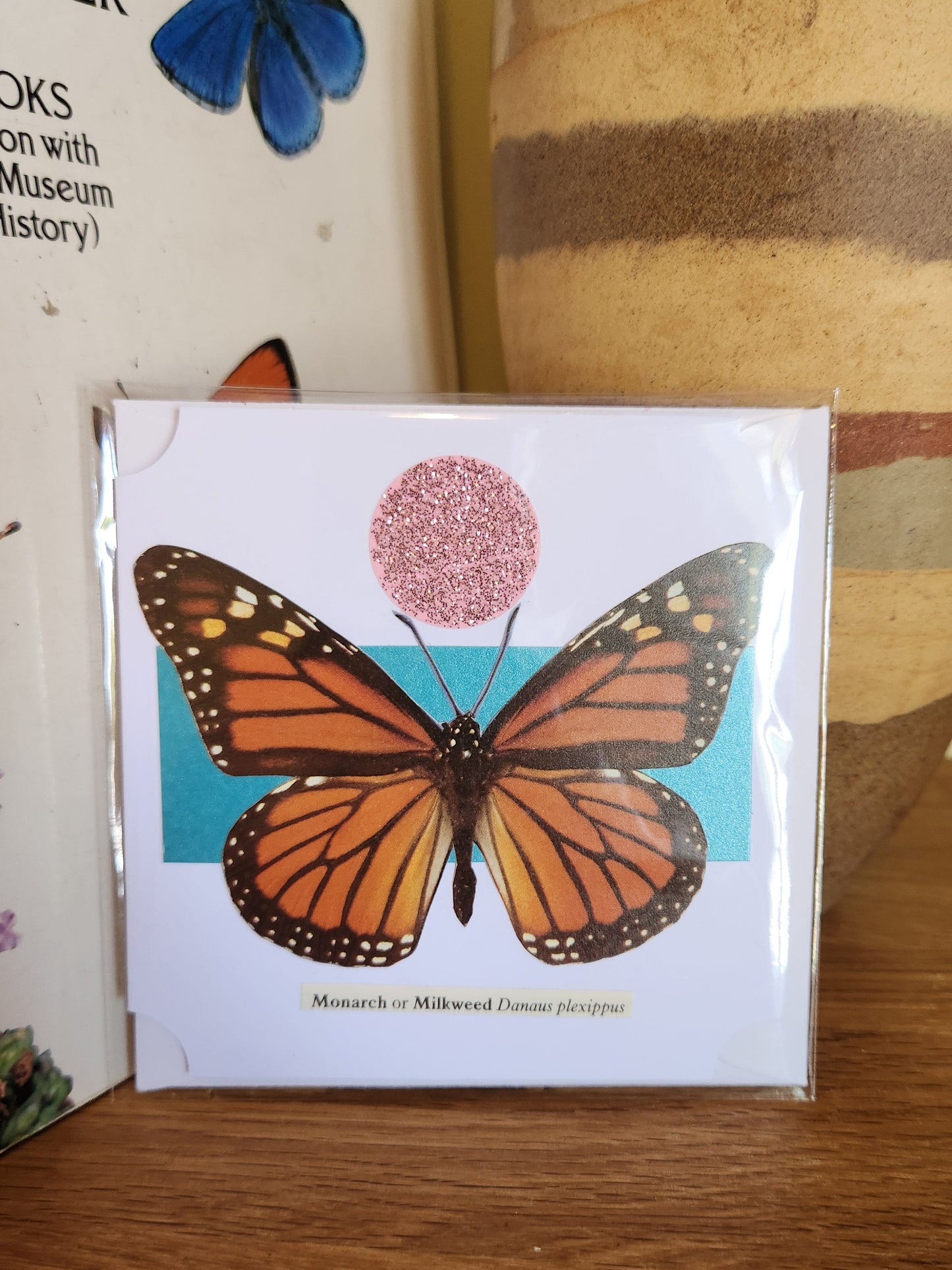A small square printed card featuring an orange monarch butterfly with a pink circular motif above it, sealed in clear plastic packaging with matching envelope. The card is placed on a wooden shelf in front of a striped ceramic vase