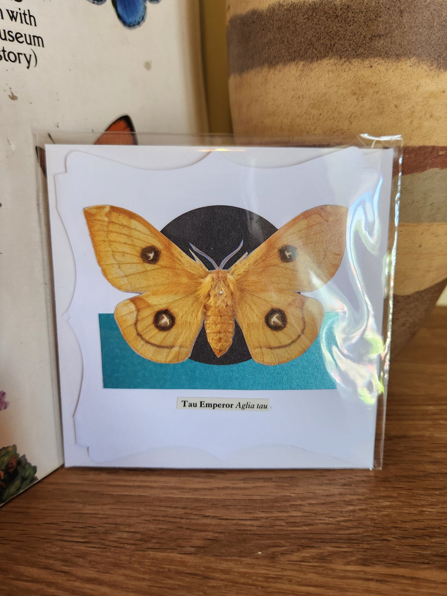 A decorative white greeting card featuring a Tau Emperor moth over a black circle and a teal rectangle, sealed in clear plastic packaging with matching envelope. The card is placed on a wooden shelf in front of a striped ceramic vase.
