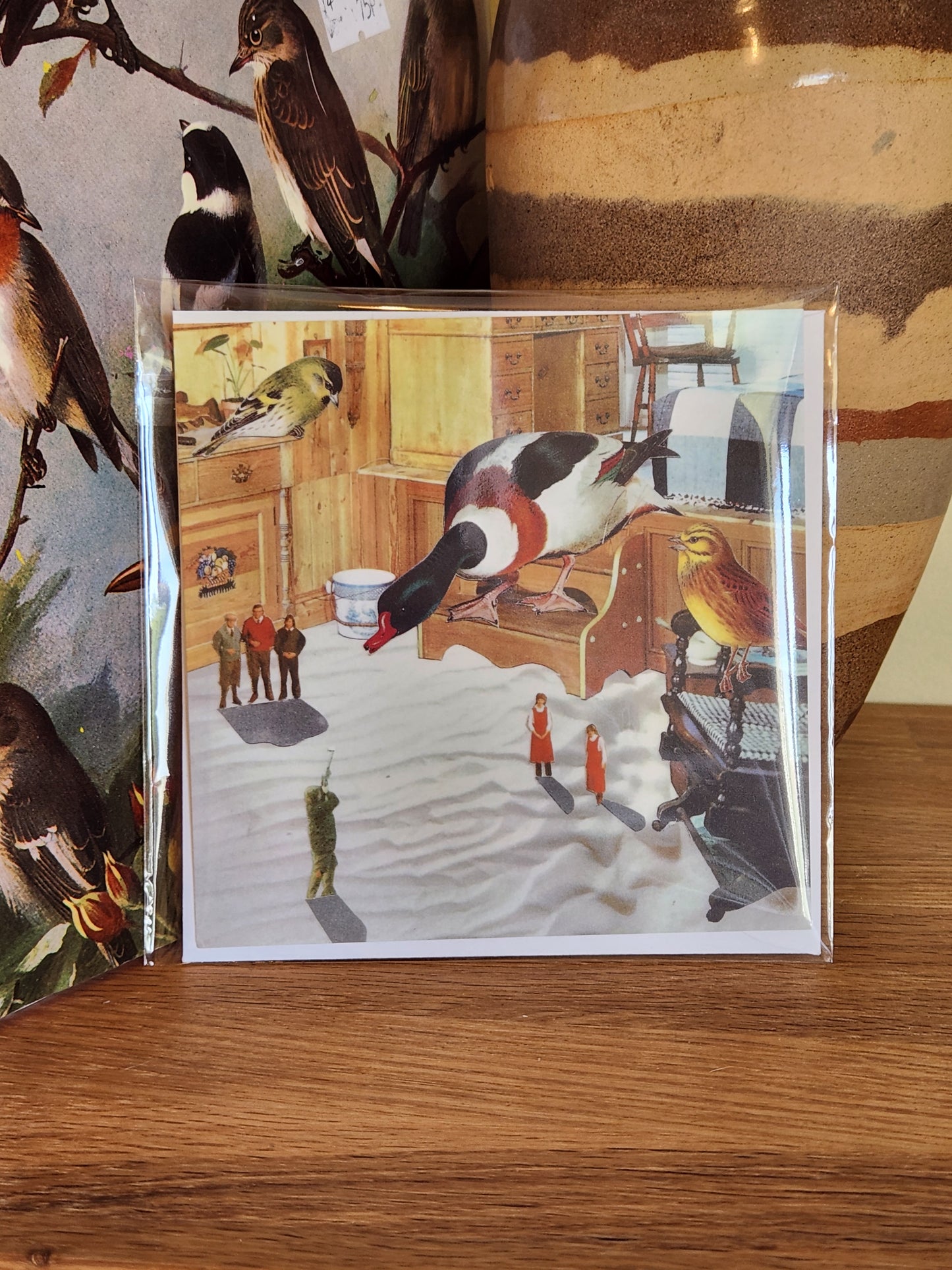 A square postcard featuring a whimsical duck hunting scene, sealed in a clear plastic sleeve with a matching envelope, and photographed on a wooden surface.