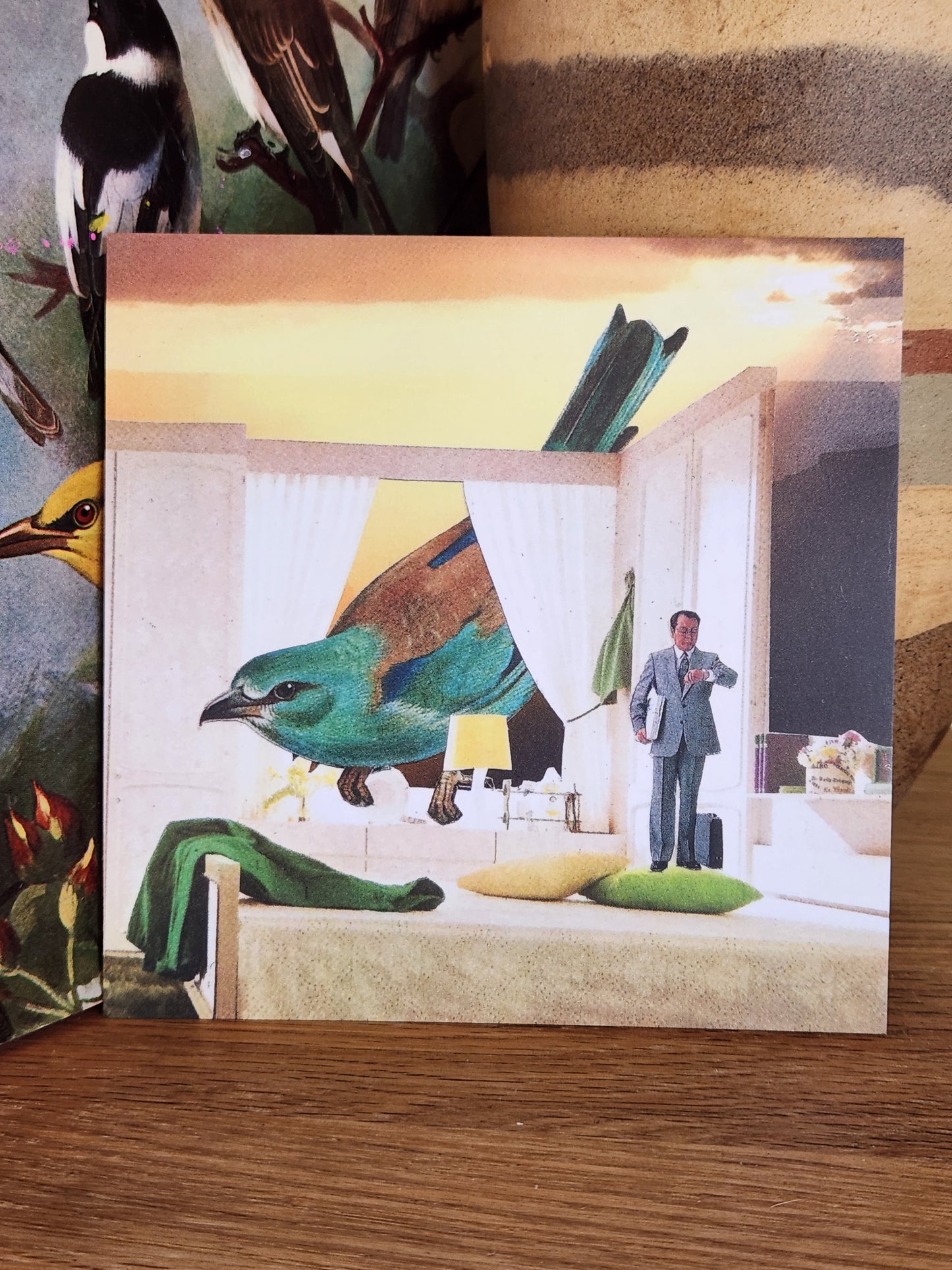 A square postcard featuring a surreal collage of a large green bird, a person in a suit, and architectural design, displayed on a wooden surface.