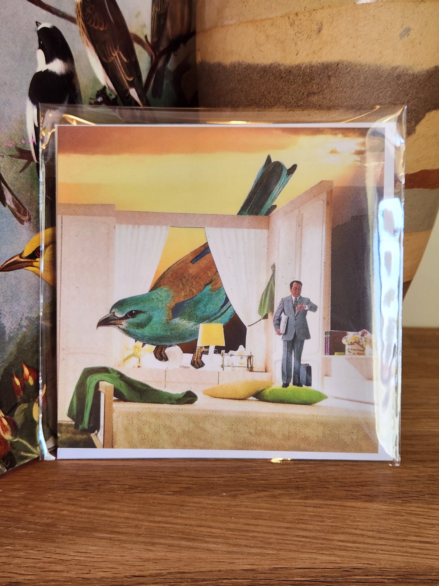 A square post card in a clear plastic sleeve with a matching envelope, showing a surreal interior scene with a large green bird-like figure and a small standing person, placed on a wooden surface.