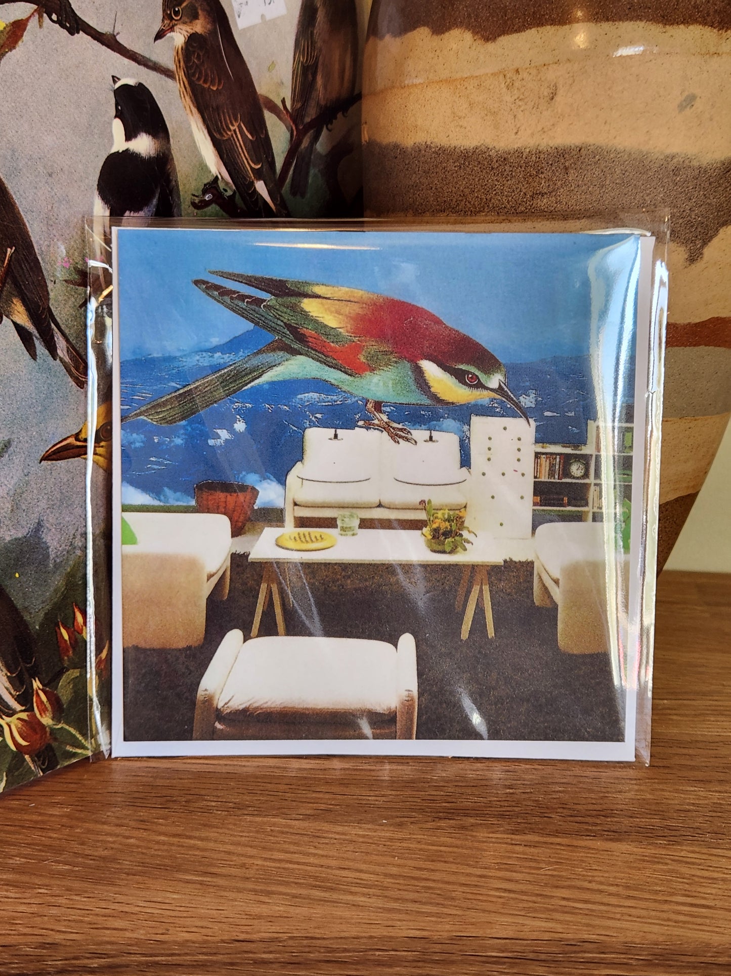 A square postcard featuring a colorful bird perched over a modern living room interior, photographed in a clear plastic sleeve with a matching envelope on a wooden surface.