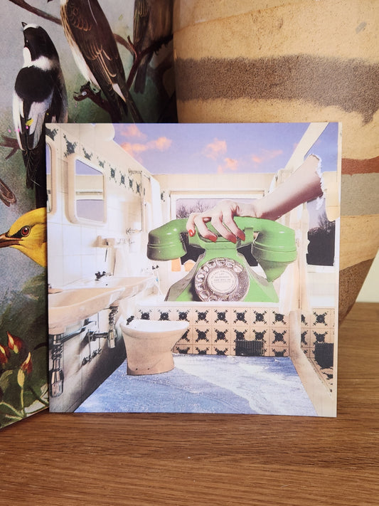 A square postcard featuring a surreal collage with a green rotary telephone, a hand, a toilet, and architectural details, photographed standing on top of a wooden surface.