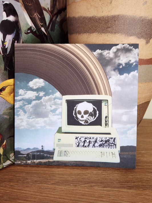 A square printed postcard placed on a wooden surface, featuring a surreal collage with a large brown arch, blue sky and clouds, a retro computer monitor with a skull graphic.