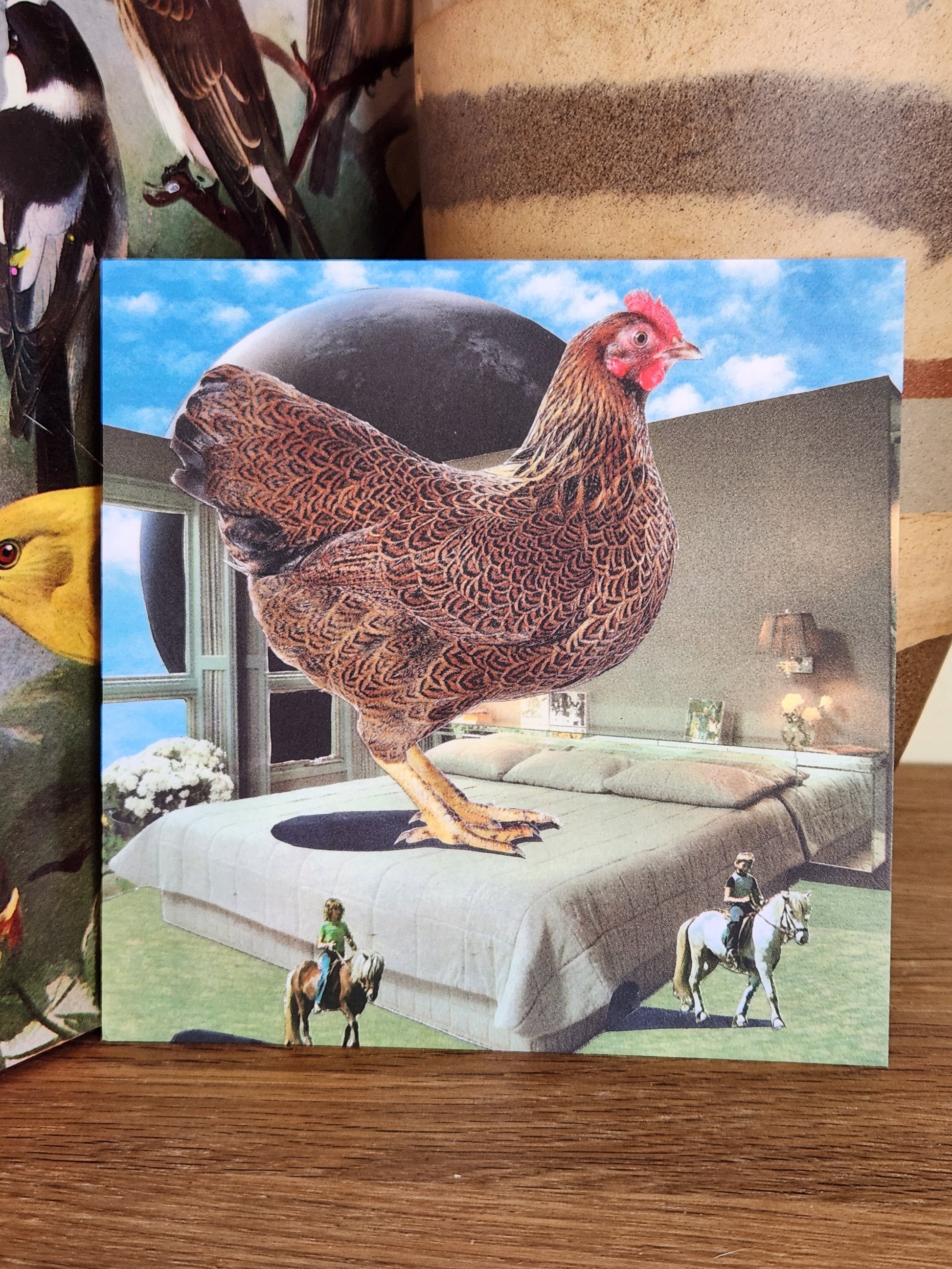 A square postcard featuring a surreal collage of a large brown chicken standing on a gray bed under a blue sky, photographed on a wooden surface with a striped ceramic vase behind it.