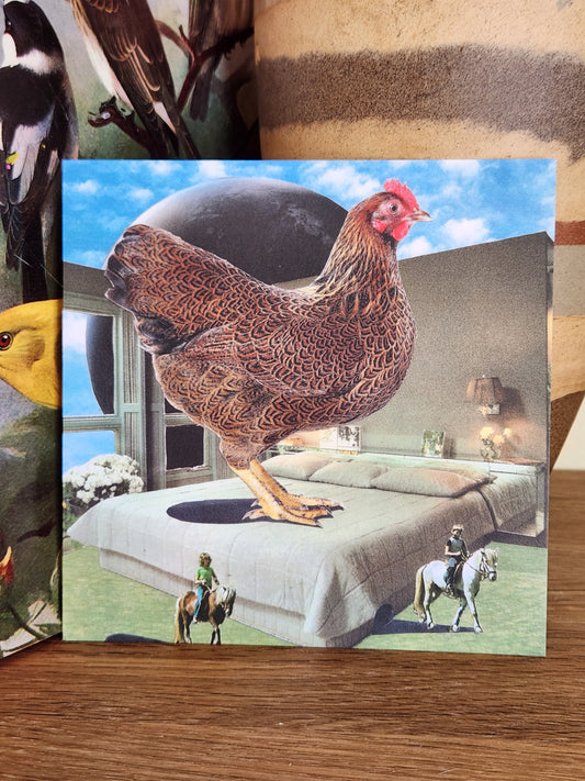 A square postcard featuring a surreal collage of a large brown chicken standing on a gray bed under a blue sky, photographed on a wooden surface with a striped ceramic vase behind it.