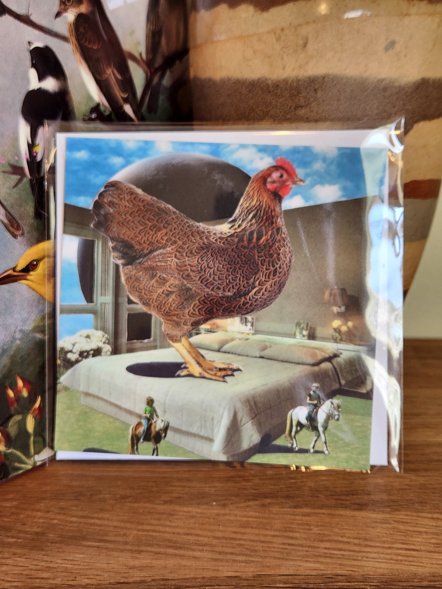 A square postcard featuring a surreal scene of a brown chicken standing on a gray bed under a blue sky, sealed in clear plastic packaging with matching envelope. The card is placed on a wooden shelf in front of a striped ceramic vase.