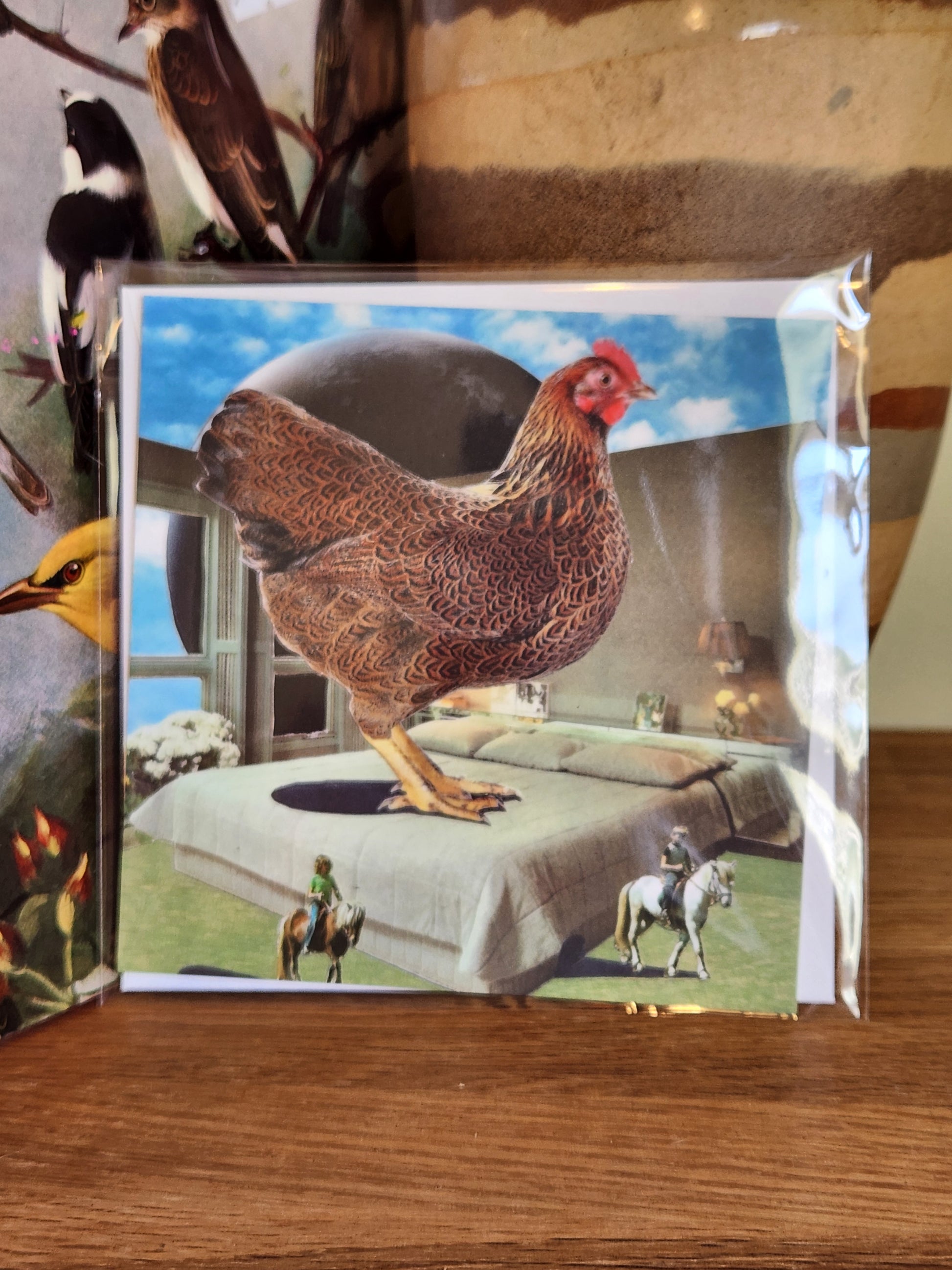 A square postcard featuring a surreal scene of a brown chicken standing on a gray bed under a blue sky, sealed in clear plastic packaging with matching envelope. The card is placed on a wooden shelf in front of a striped ceramic vase.
