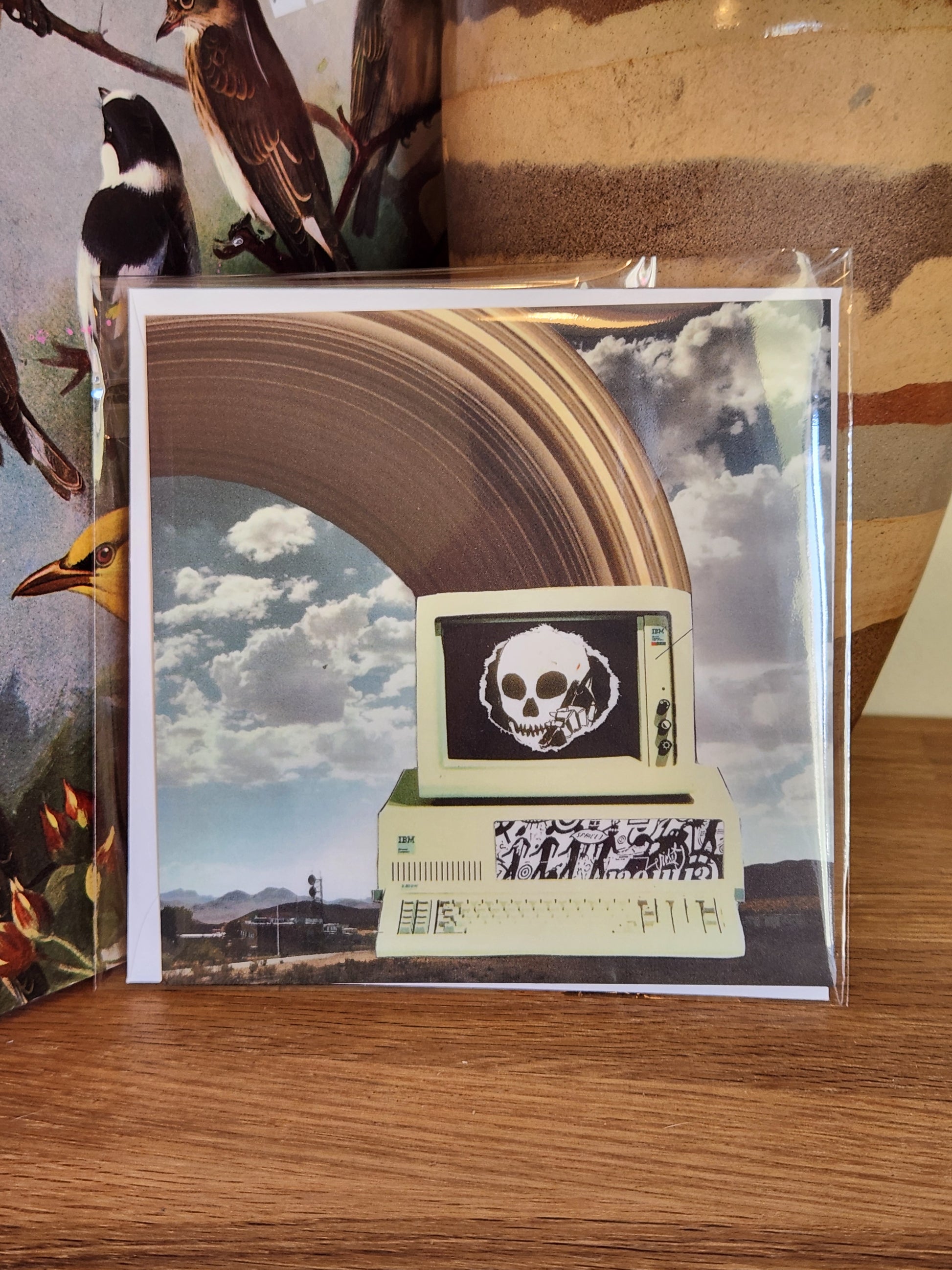 A square printed postcard with matching envelope in a clear plastic sleeve, placed on a wooden surface. The artwork shows a surreal collage with a large brown arch, cloudy sky, a skull inside a monitor-like frame, and a graffiti-style graphic.