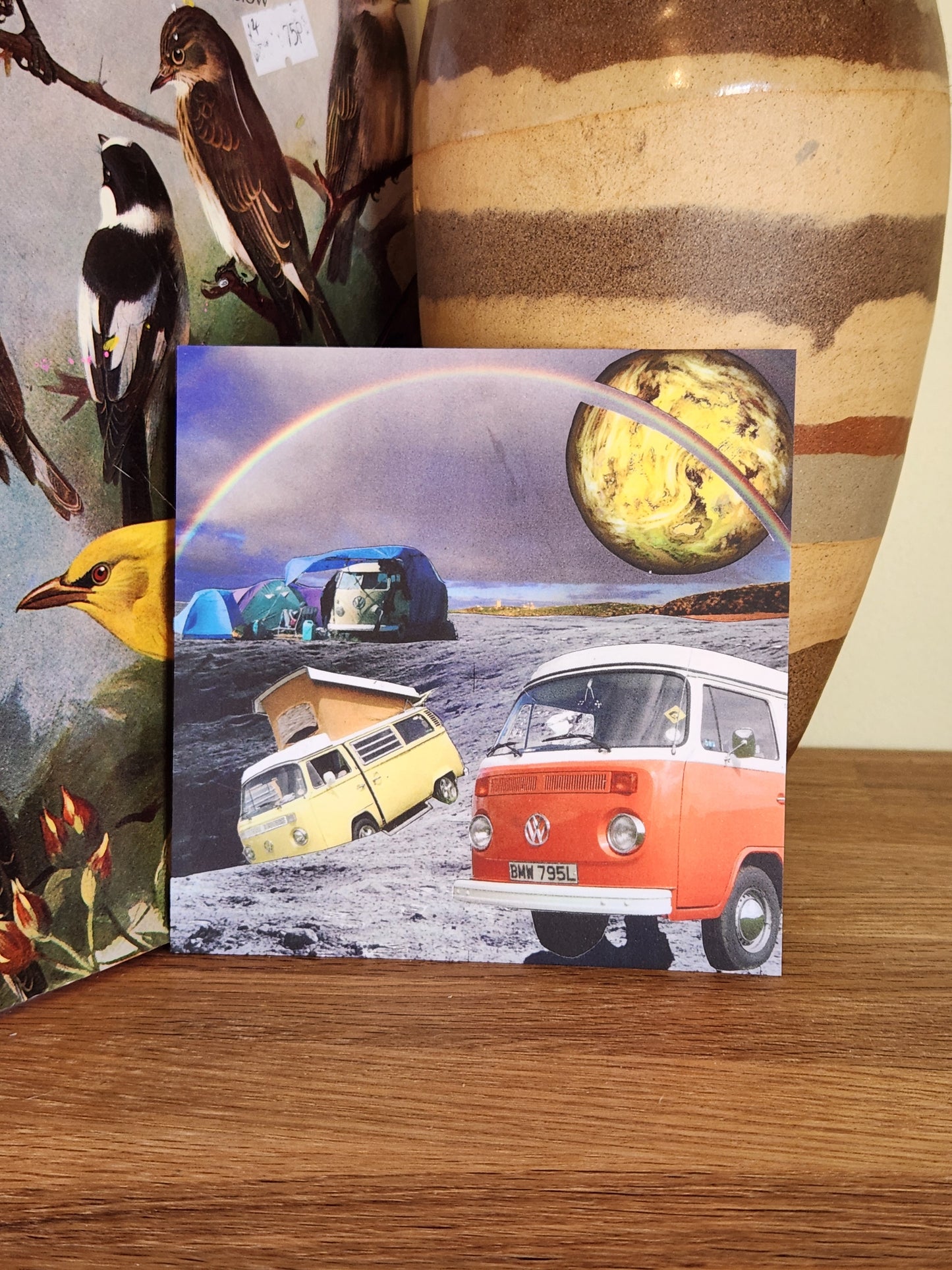 A square postcard featuring a surreal collage of vintage VW camper vans, a rainbow, and a planet, displayed on a wooden surface next to a large ceramic vase.