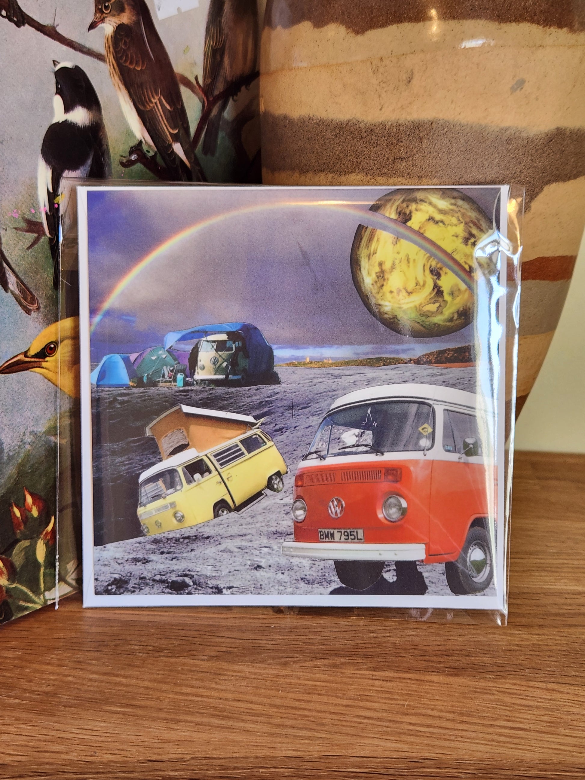 A square postcard featuring a surreal collage of vintage VW camper vans, a rainbow, and a yellow-green planet, sealed in clear plastic packaging with matching envelope. The card is placed on a wooden shelf in front of a striped ceramic vase.